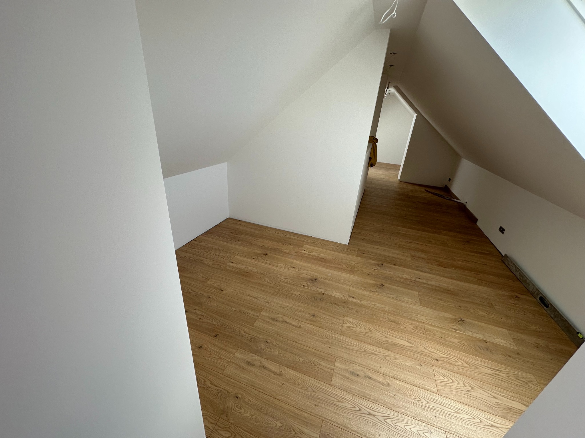 Poddasze z nową drewnianą podłogą i świeżo pomalowanymi na biało ścianami, widoczny fragment poziomicy przy ścianie oraz wieszak na ubrania w tle.