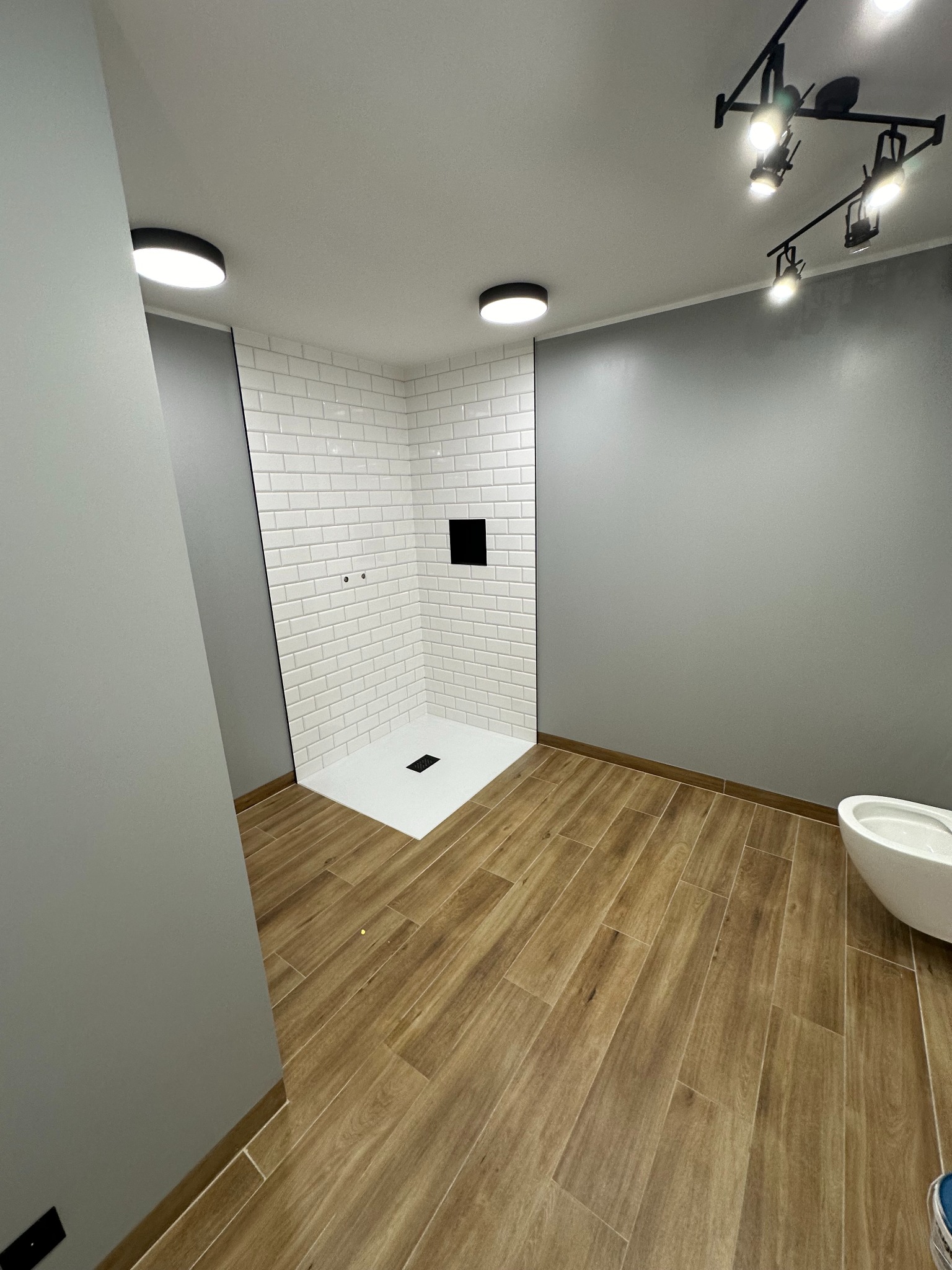 Wykończona łazienka w Białymstoku z kabiną prysznicową wyłożoną białymi, prostokątnymi płytkami, drewnopodobną podłogą i szarą ścianą, oświetlona punktowymi lampami.