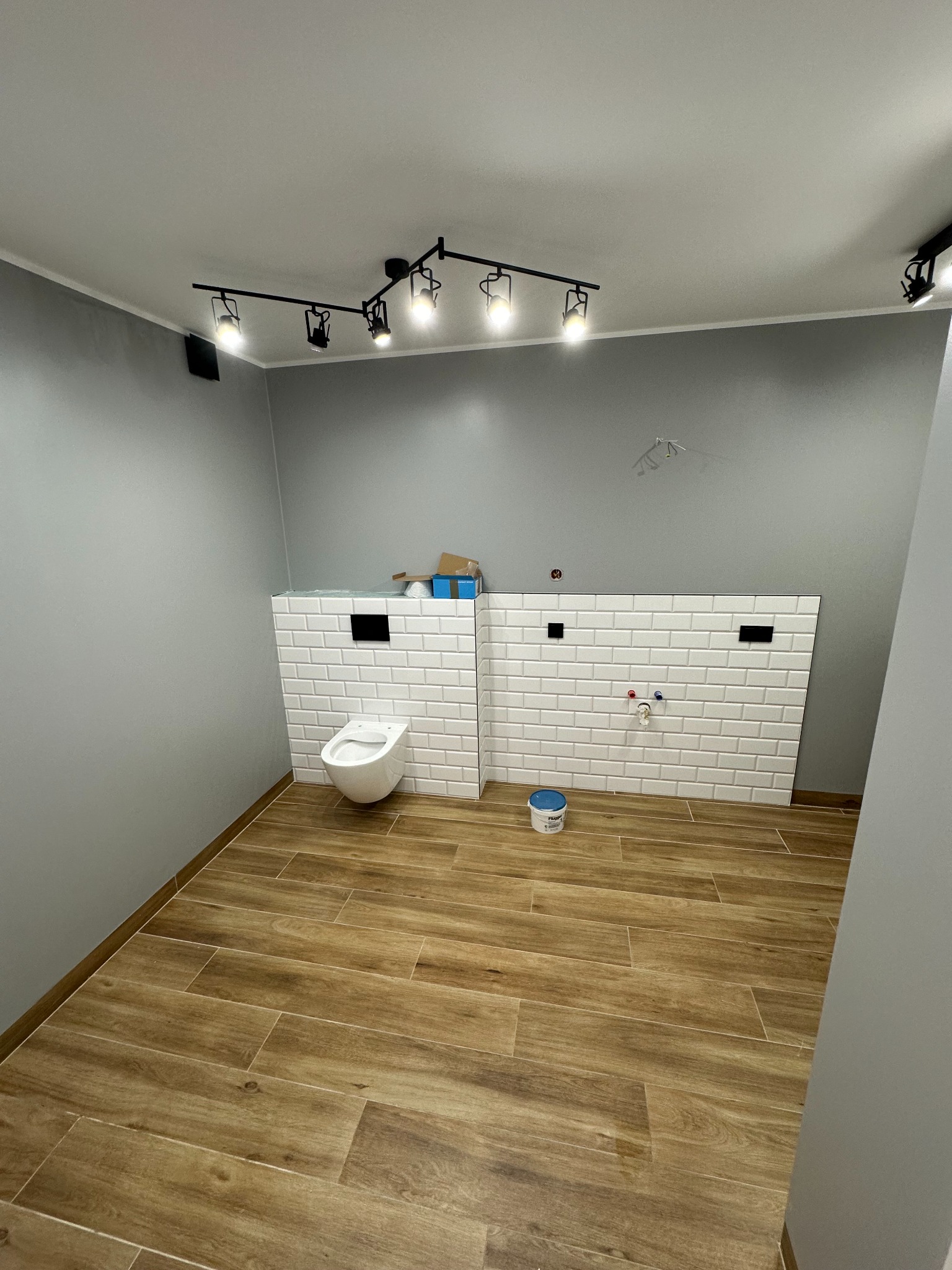 Wnętrze łazienki w trakcie wykończenia: drewnopodobna podłoga, szara ściana, zabudowa stelaża WC z białych, prostokątnych płytek, wisząca toaleta i czarne oświetlenie sufitowe.