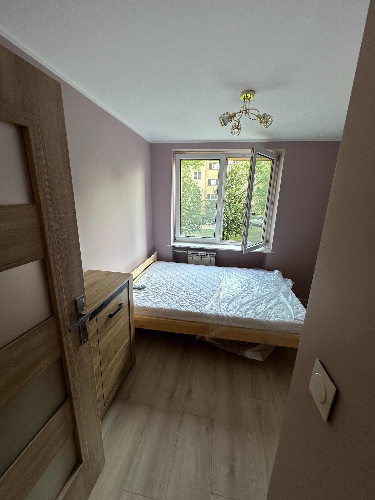 Pokój z łóżkiem zabezpieczonym folią malarską, ściany pomalowane na liliowo, widoczne drewniane panele podłogowe i fragment drewnianej komody z drzwiami.