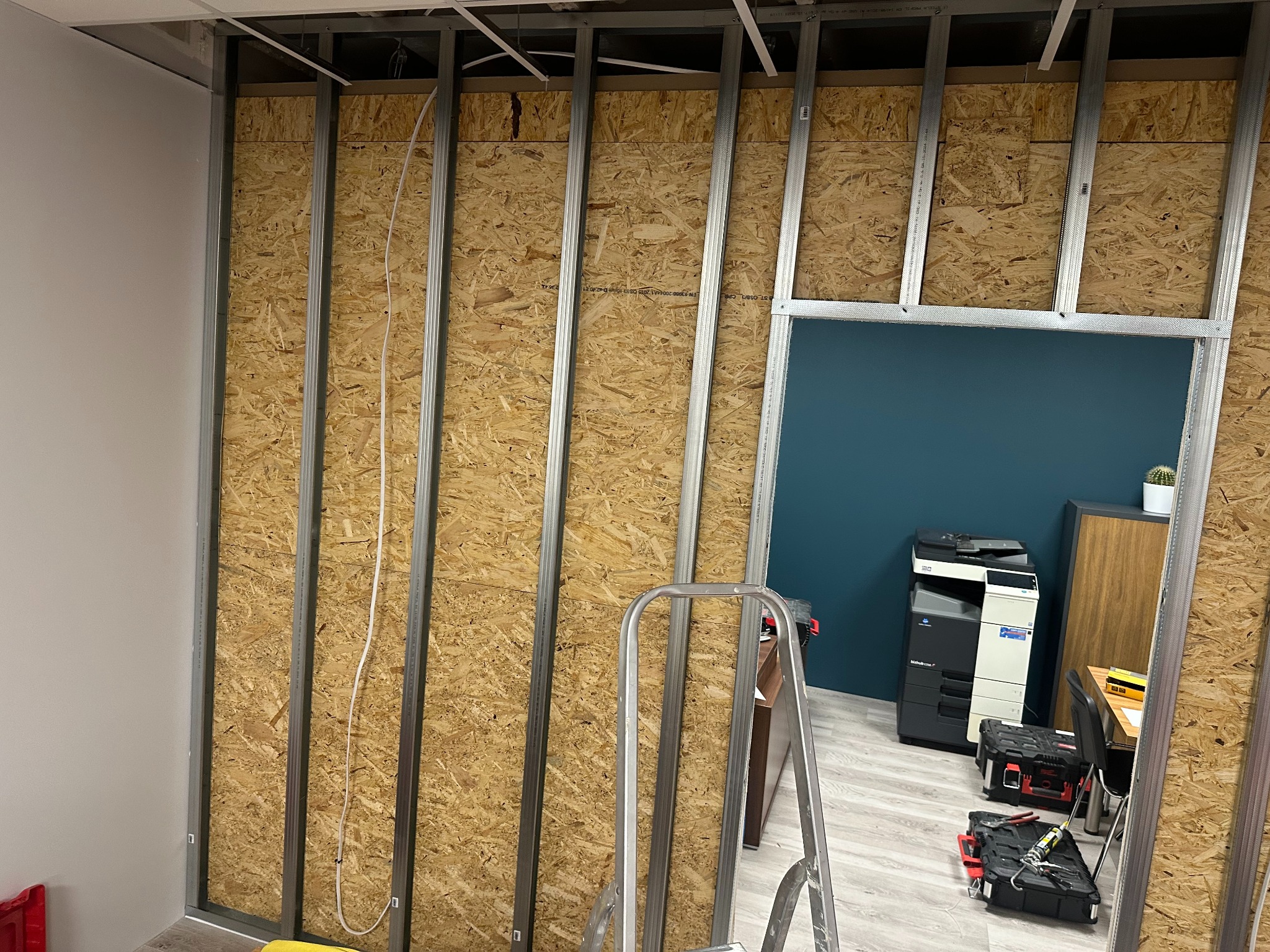 Stelaż z profili stalowych i płyty OSB na ścianie działowej w biurze, widoczne przewody elektryczne, drabina, biurko, drukarka i skrzynki narzędziowe w tle.