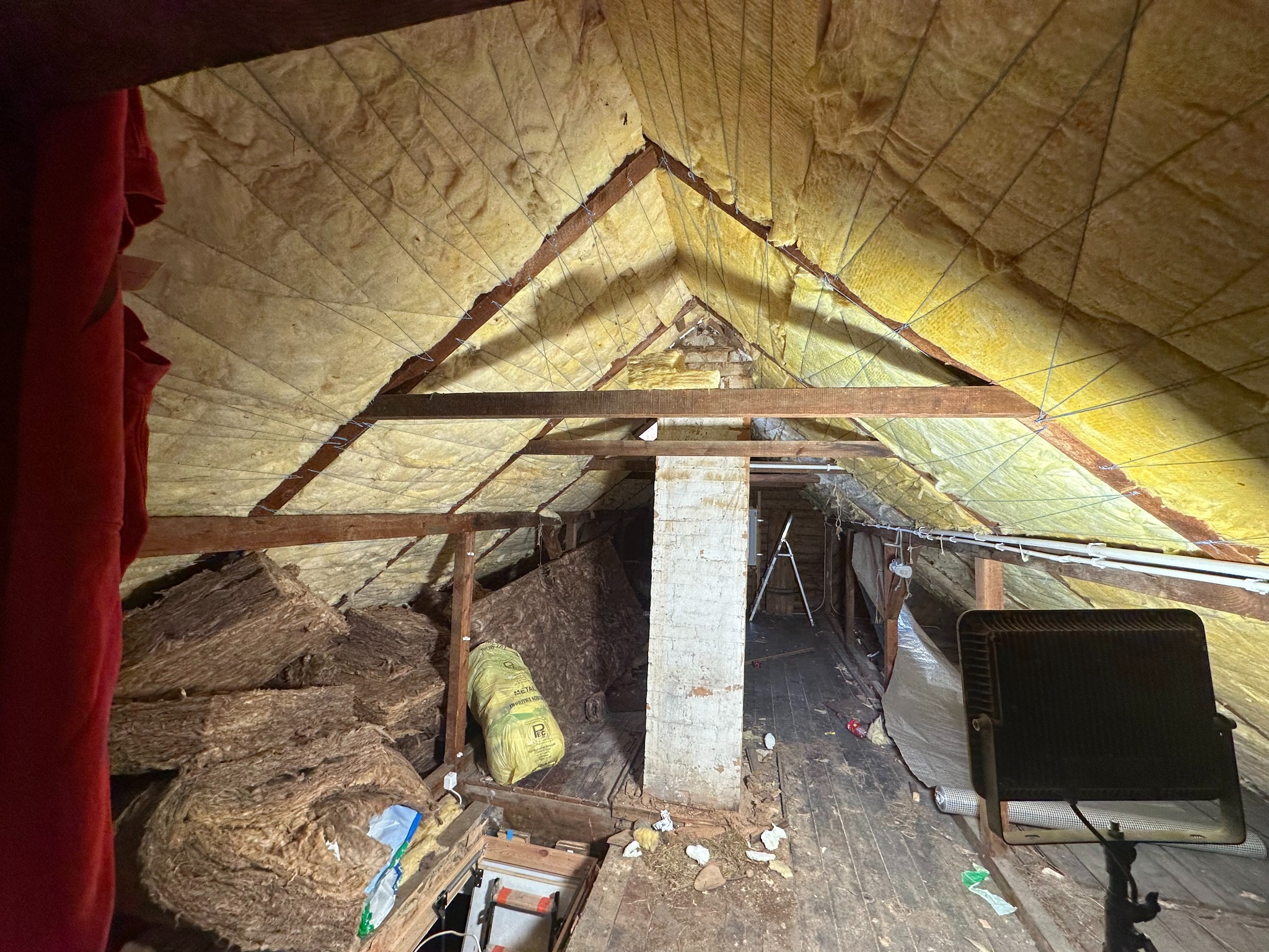 Poddasze z drewnianą konstrukcją dachu, ocieplone wełną mineralną, widoczne fragmenty instalacji elektrycznej, sterta wełny w rogu oraz lampa halogenowa na podłodze.