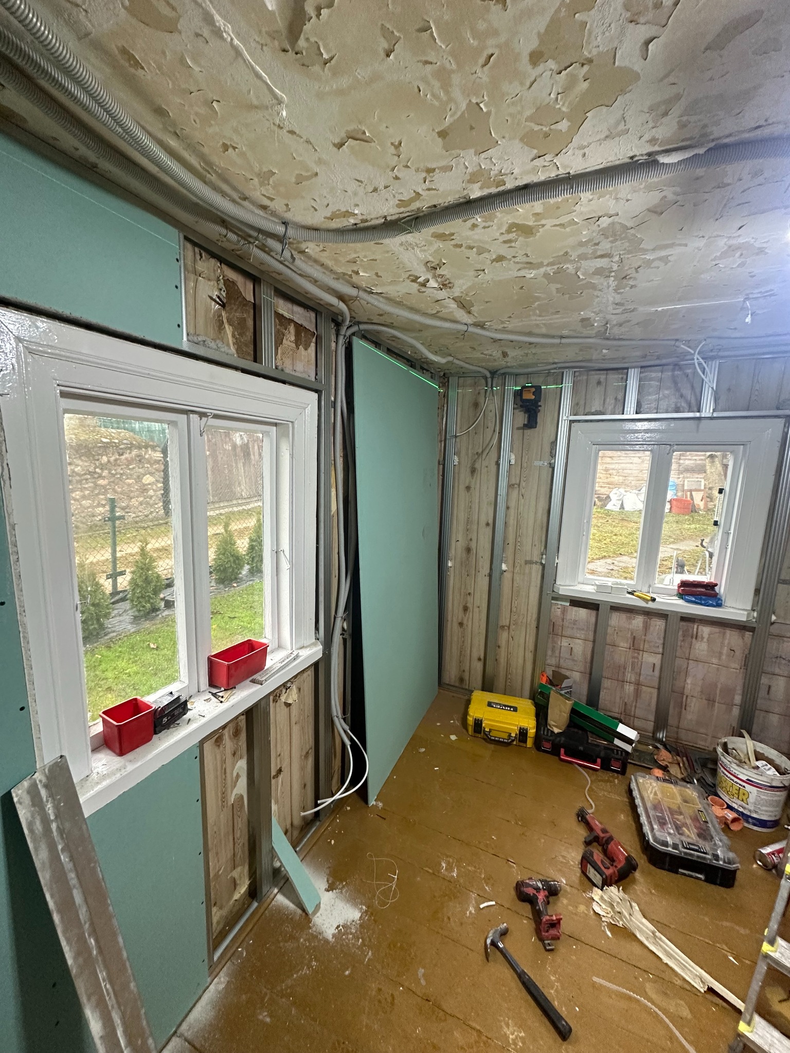 Wnętrze w trakcie remontu: ściany z metalowym stelażem, częściowo obłożone płytami gipsowo-kartonowymi w kolorze zielonym, widoczne instalacje elektryczne pod sufitem z odspojoną farbą, narzędzia...