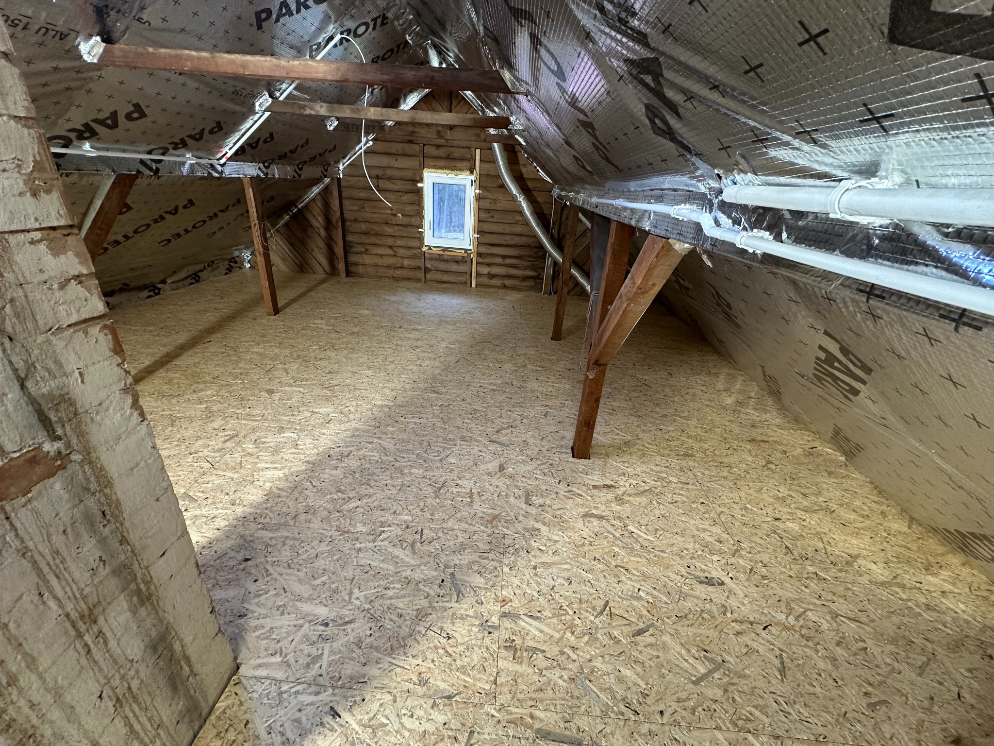 Poddasze z drewnianą konstrukcją, ocieplone folią paroizolacyjną z widocznymi rurami wentylacyjnymi pod sufitem i podłogą wyłożoną płytami OSB, małe okno w tylnej ścianie szczytowej.