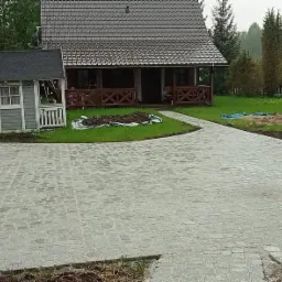 Ułożona szara kostka brukowa na podjeździe przed domem z brązową werandą i domkiem dla dzieci po lewej, widoczne ślady po pracach ziemnych.