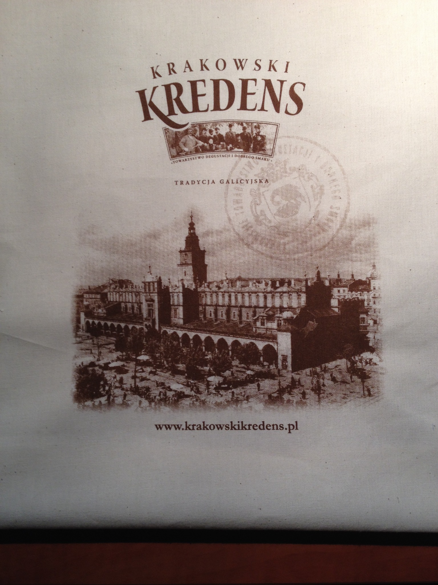 Beżowa torba bawełniana z brązowym nadrukiem przedstawiającym Sukiennice w Krakowie i logo 'Krakowski Kredens'.