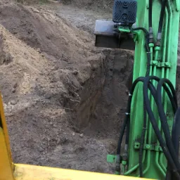 Zielona koparka z czarnymi przewodami hydraulicznymi częściowo widoczna z kabiny operatora, obok wykopu w ziemi z widocznymi korzeniami.