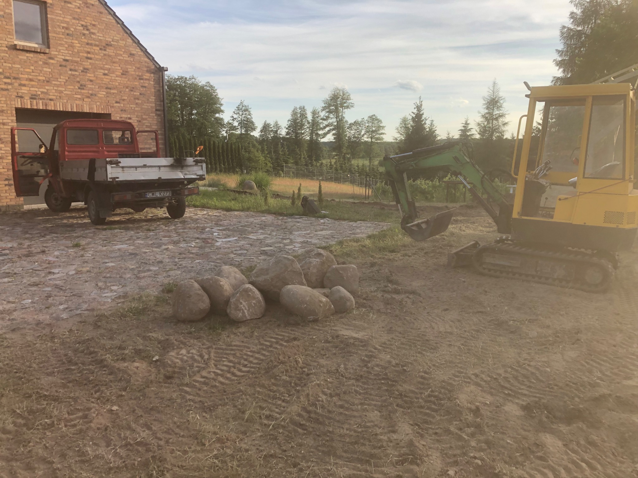 Żółta mini koparka na nierównym terenie obok sterty dużych kamieni, w tle czerwono-biała ciężarówka i ceglany budynek gospodarczy, widoczne ślady opon na ziemi.