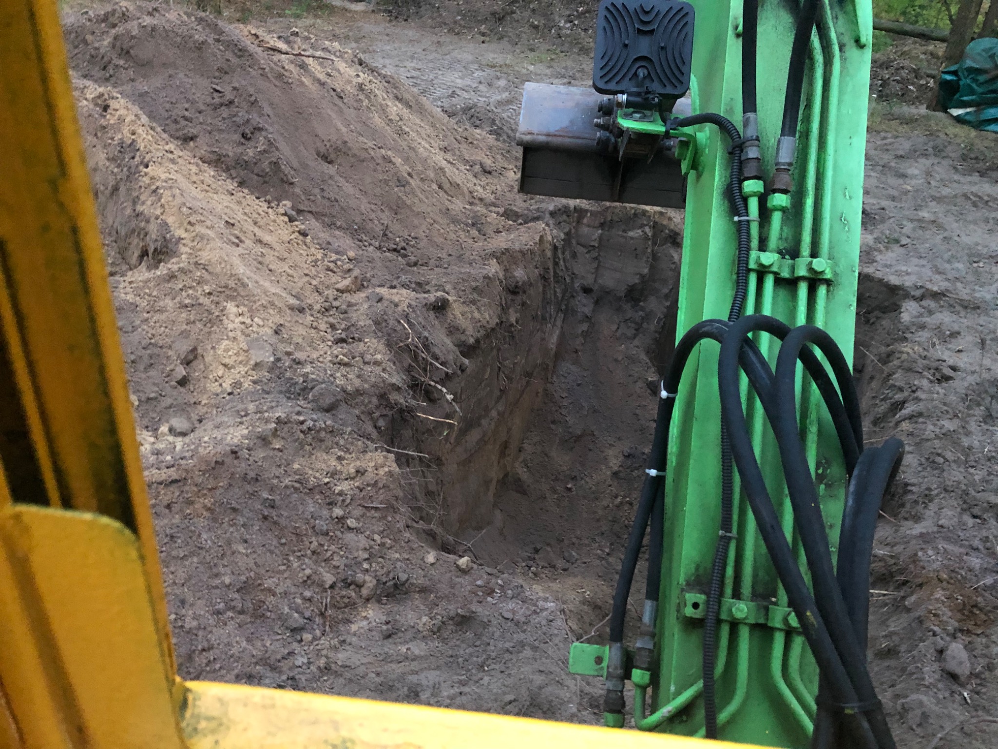 Zielona koparka z czarnymi przewodami hydraulicznymi częściowo widoczna z kabiny operatora, obok wykopu w ziemi z widocznymi korzeniami.