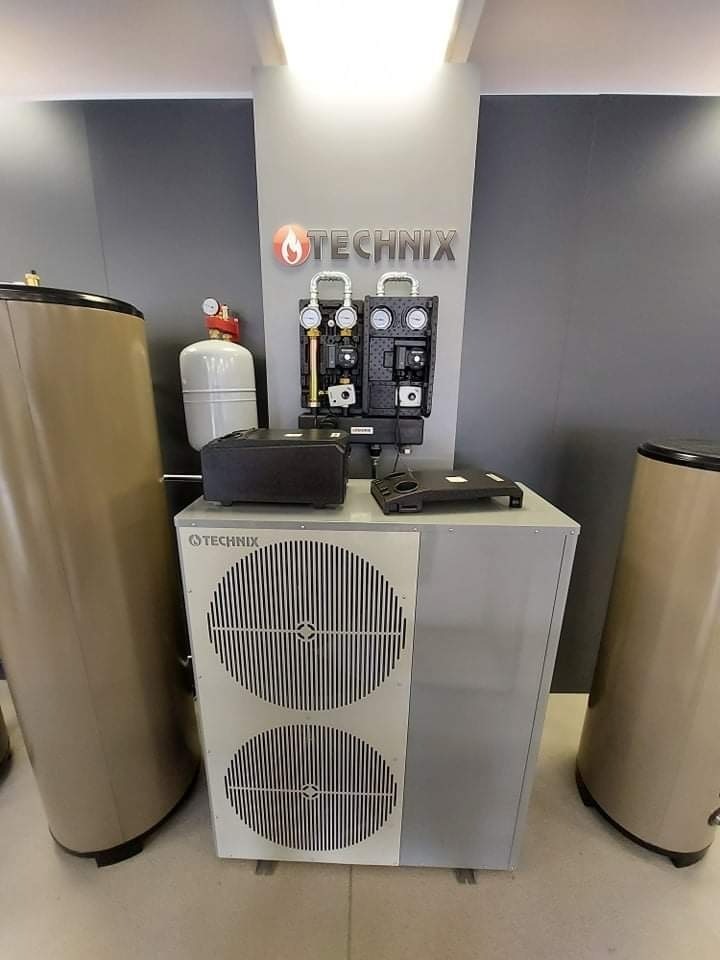 Prezentacja urządzenia grzewczego marki Technix w Poznaniu: szara obudowa z dwoma okrągłymi wlotami powietrza, zbiorniki buforowe po bokach, manometry i sterowniki na górze, logo firmy na ścianie...