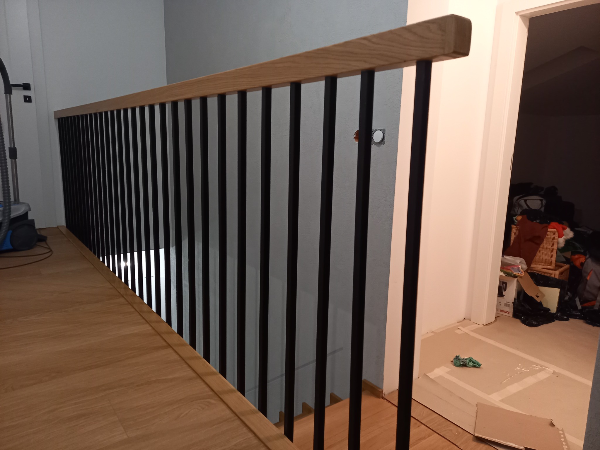 Nowoczesna balustrada z czarnymi, pionowymi słupkami i drewnianym blatem, widoczna z góry, z podłogą w odcieniach drewna i fragmentem pomieszczenia w trakcie remontu.