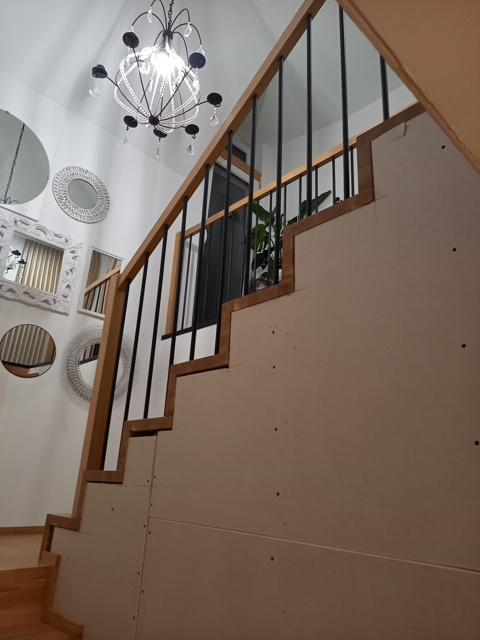 Wnętrze z drewnianymi schodami w trakcie montażu, widoczne niedokończone płyty gipsowo-kartonowe, czarne metalowe balustrady i ozdobne lustra na ścianie.