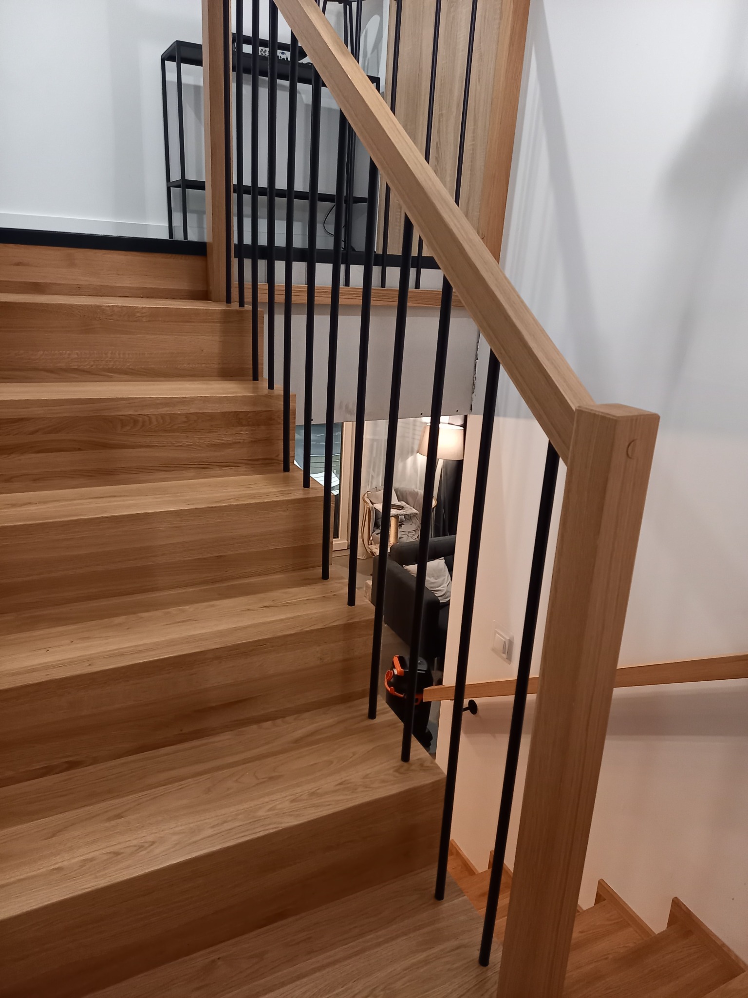 Jasne, drewniane stopnie schodów z czarnymi, metalowymi balustradami, widok z góry na dół, z fragmentem salonu w tle.
