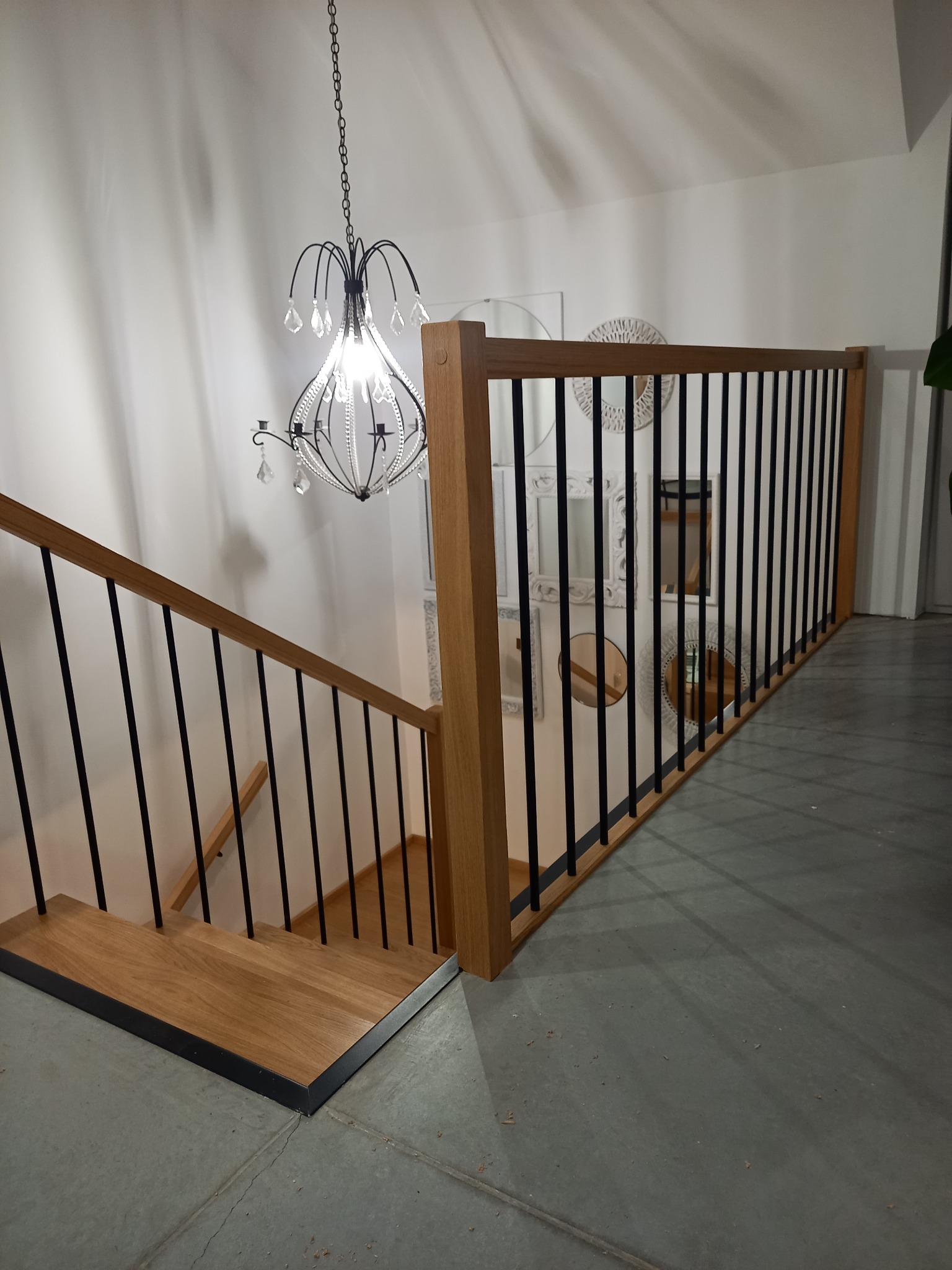 Nowoczesne schody z drewnianymi stopniami i czarnymi metalowymi balustradami, oświetlone dekoracyjnym żyrandolem w stylu glamour, na tle ściany z lustrami w ozdobnych ramach.