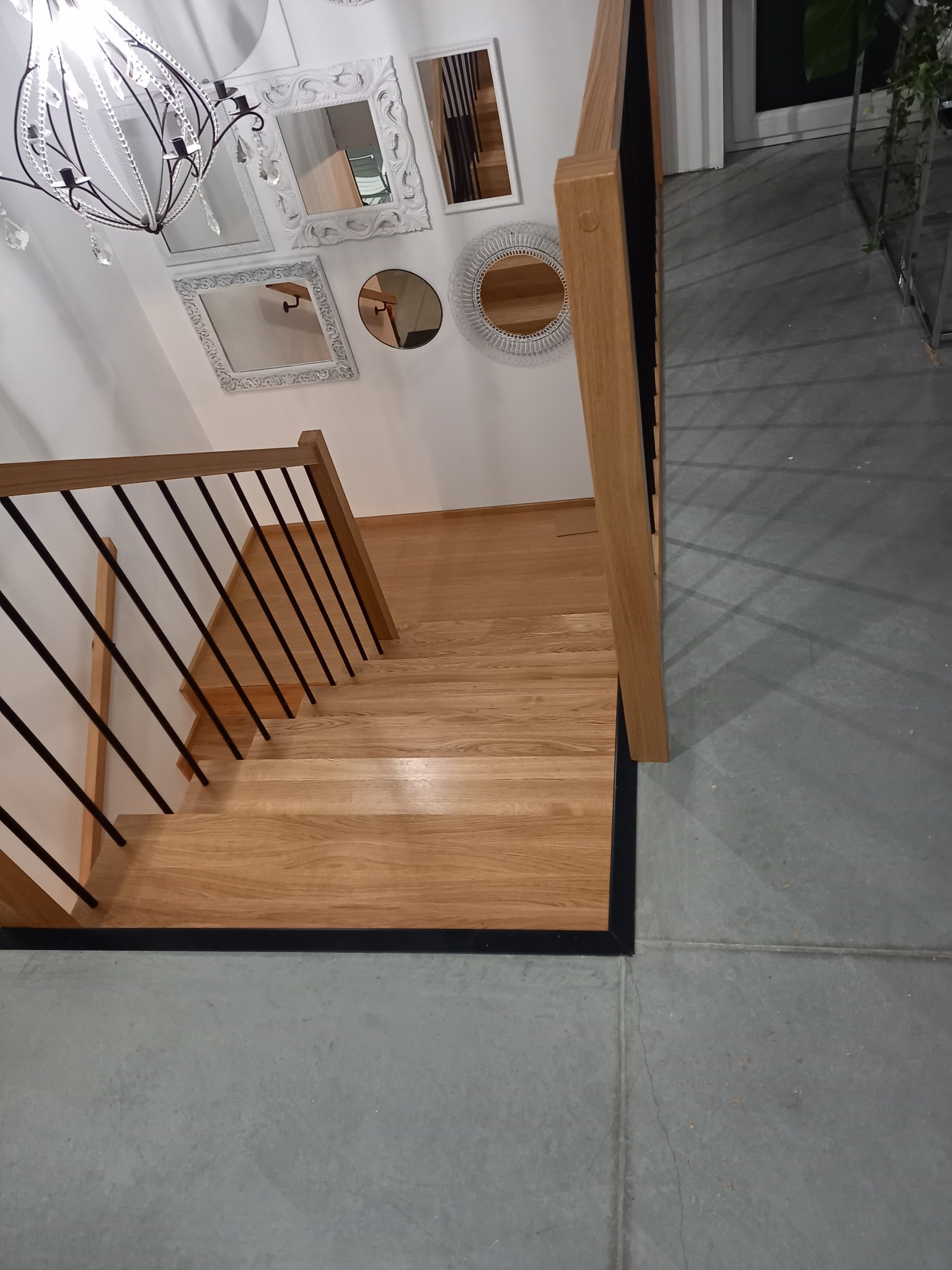 Widok z góry na jasne, dębowe schody z czarnymi, metalowymi balustradami w Częstochowie, oświetlone wiszącym żyrandolem i otoczone dekoracyjnymi lustrami.