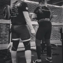 Dwóch mężczyzn w odzieży sportowej, jeden z pasem do podnoszenia ciężarów, przygotowuje się do podnoszenia sztangi w siłowni z ceglaną ścianą w tle.