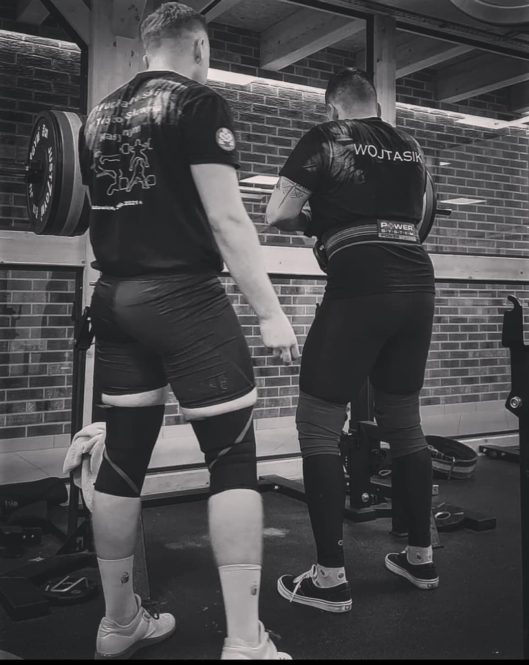Dwóch mężczyzn w odzieży sportowej, jeden z pasem do podnoszenia ciężarów, przygotowuje się do podnoszenia sztangi w siłowni z ceglaną ścianą w tle.