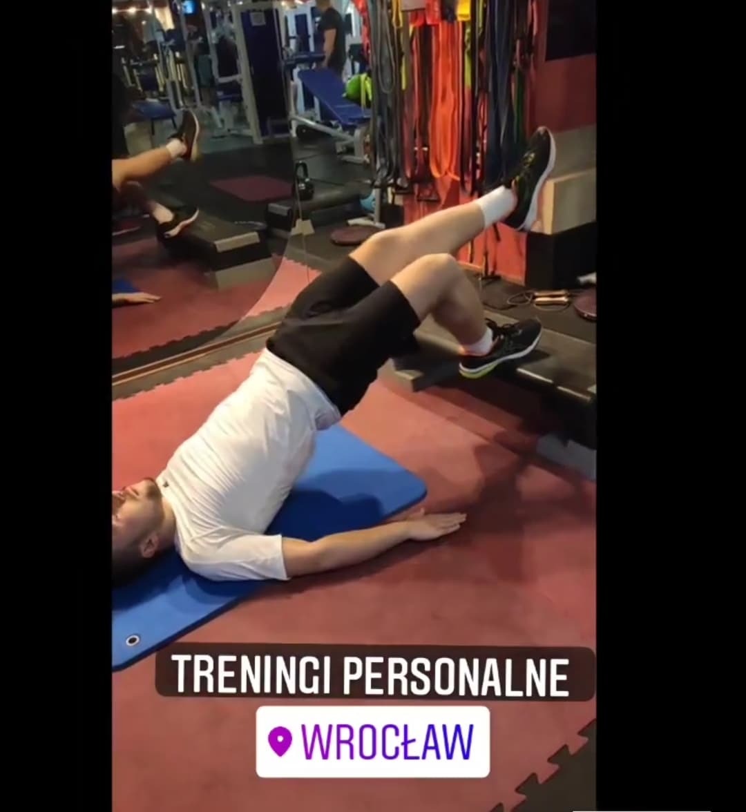 Mężczyzna w białej koszulce wykonuje ćwiczenie unoszenia bioder z jedną nogą opartą na ławce w siłowni, leżąc na niebieskiej macie, w tle widoczne wyposażenie siłowni i lustro.