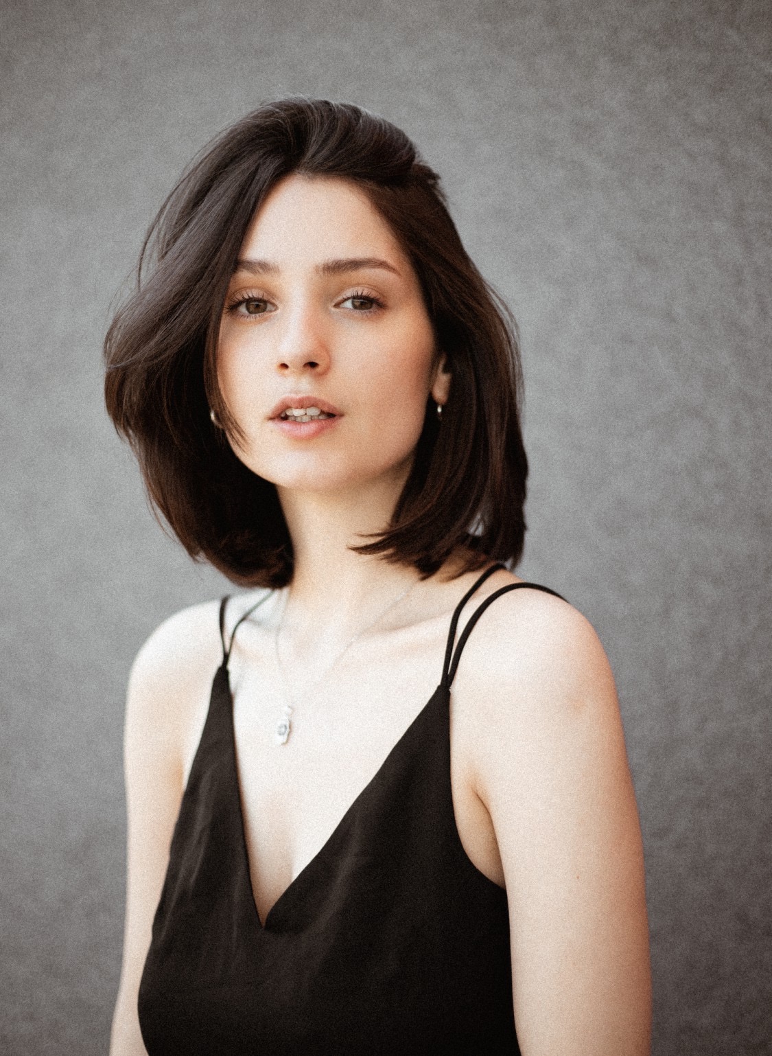 Portret młodej kobiety z krótkimi, ciemnymi włosami, w czarnym topie na ramiączkach i srebrnym naszyjniku, na tle szarej ściany.