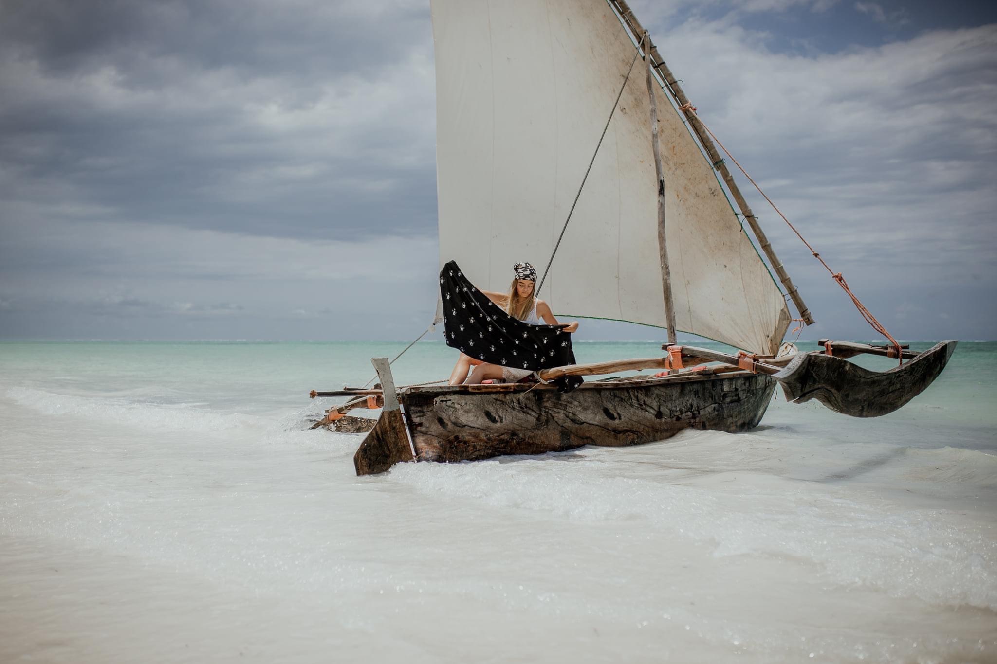 Kobieta siedzi w tradycyjnej drewnianej łodzi żaglowej na płytkiej, turkusowej wodzie, trzymając czarny szal z białym wzorem na tle pochmurnego nieba.