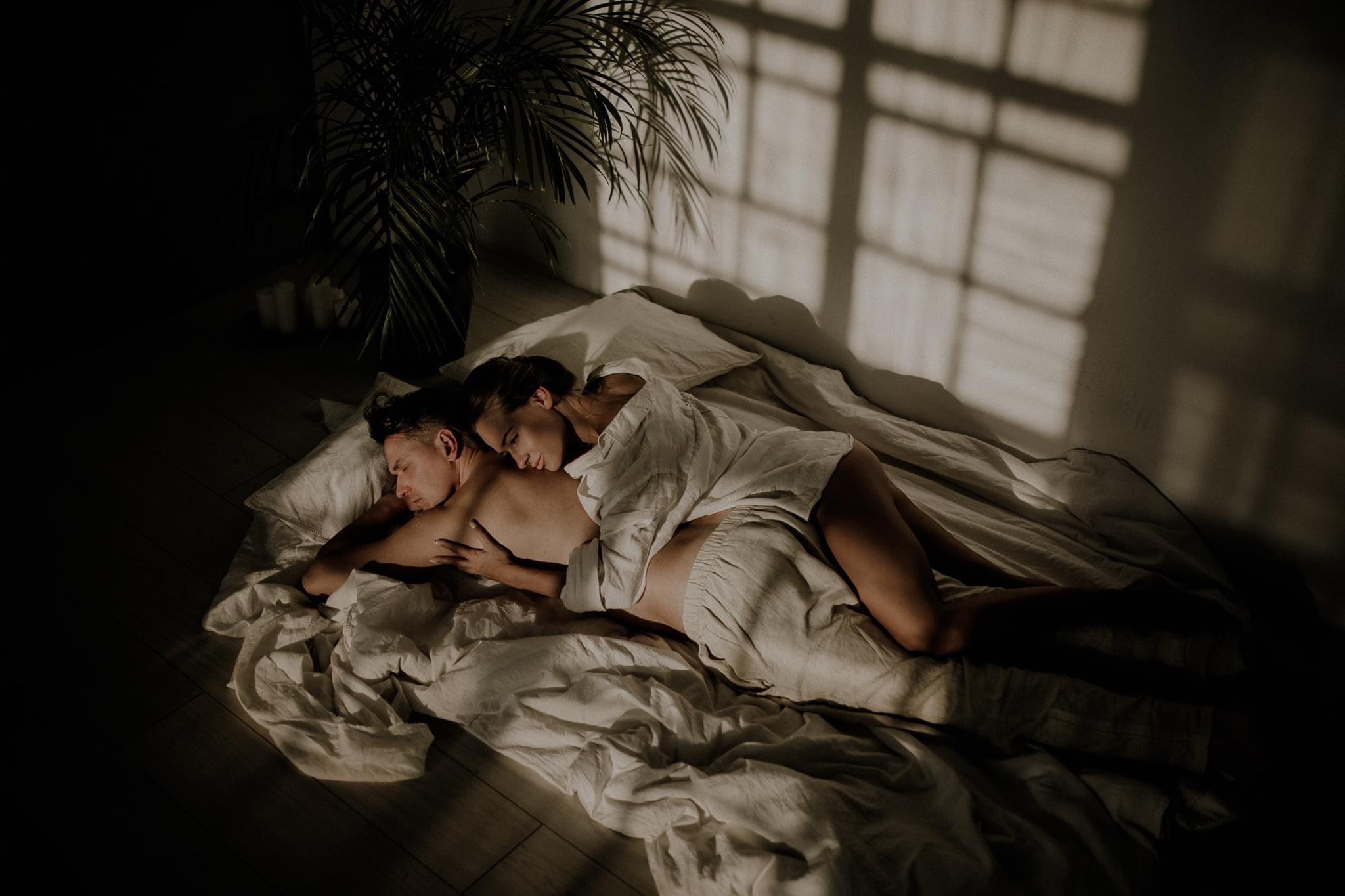 Para zakochanych we Wrocławiu, leżąca na łóżku w pościeli w odcieniach bieli, wpadające przez okno światło rzuca cienie na ścianę, obok stoi roślina doniczkowa.