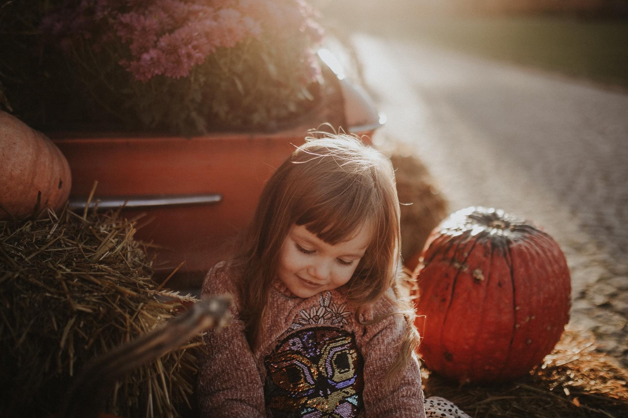 Mała dziewczynka w swetrze z cekinami siedzi obok dyni i siana, z tyłu czerwony samochód z kwiatami, ciepłe światło słoneczne.