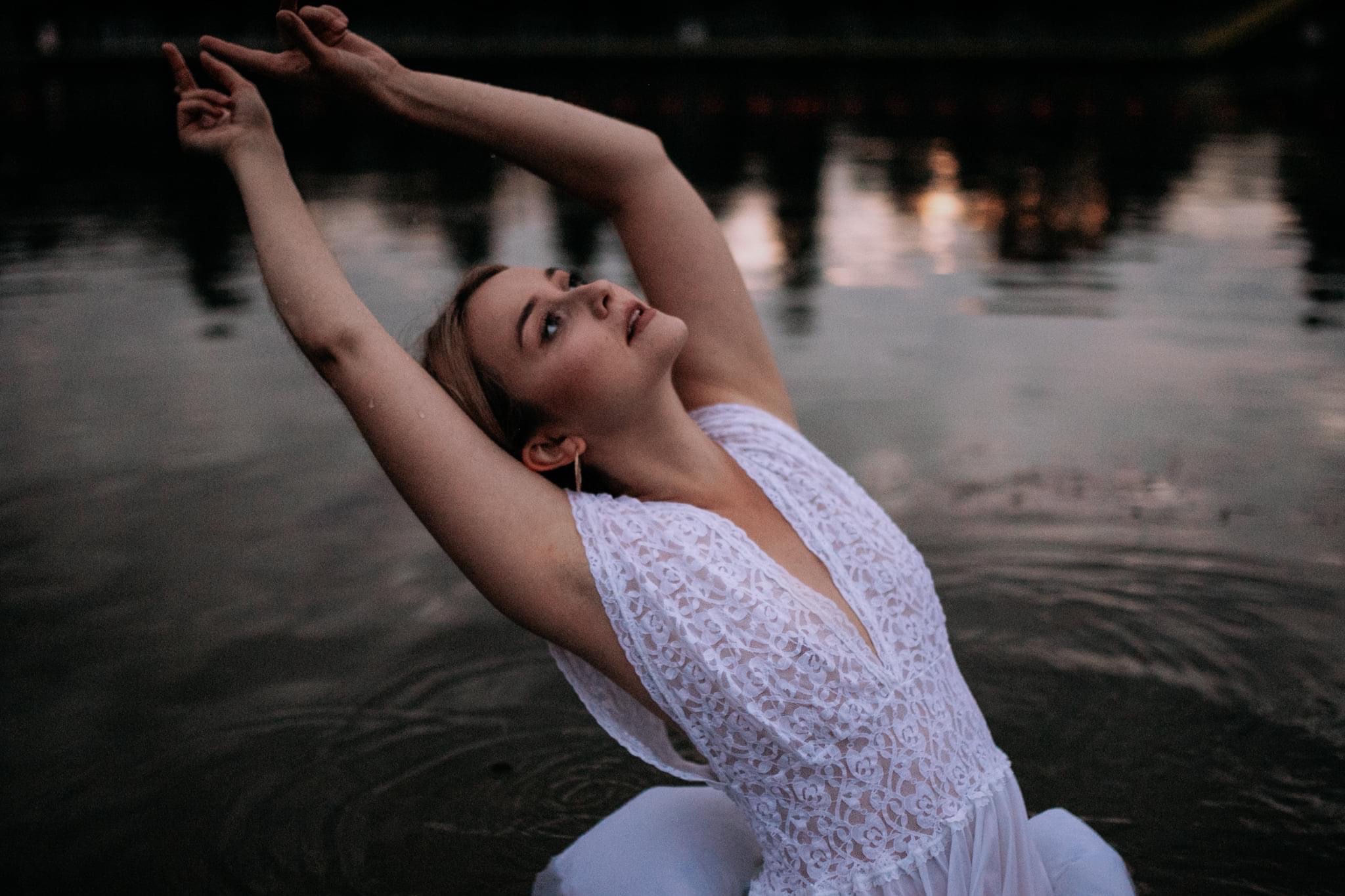 Młoda kobieta w koronkowej sukience, stojąca w wodzie, z uniesionymi rękami i spojrzeniem skierowanym ku górze, na tle rozmytego krajobrazu z zachodzącym słońcem.