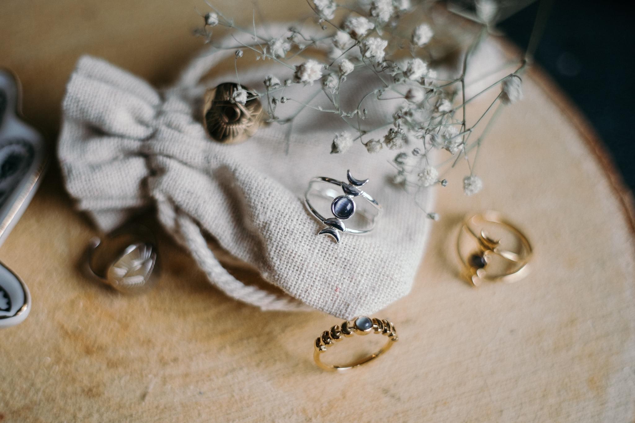 Delikatne, srebrne i złote pierścionki z motywem księżyca i okrągłym kamieniem, leżące na drewnianej powierzchni obok lnianego woreczka i gałązek gipsówki.