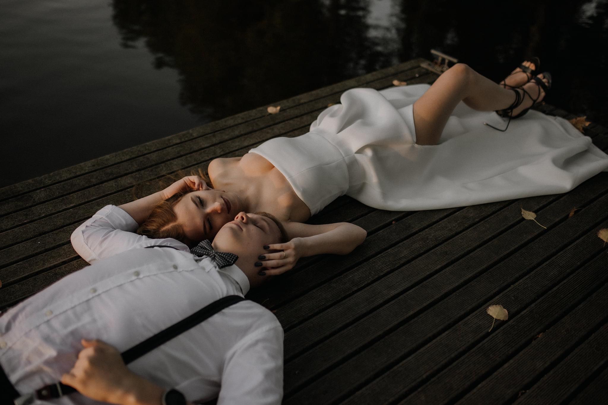 Para narzeczonych leżąca na drewnianym pomoście nad wodą: panna młoda w białej sukni, pan młody w białej koszuli z muchą i szelkami, oboje z zamkniętymi oczami, w delikatnym oświetleniu.