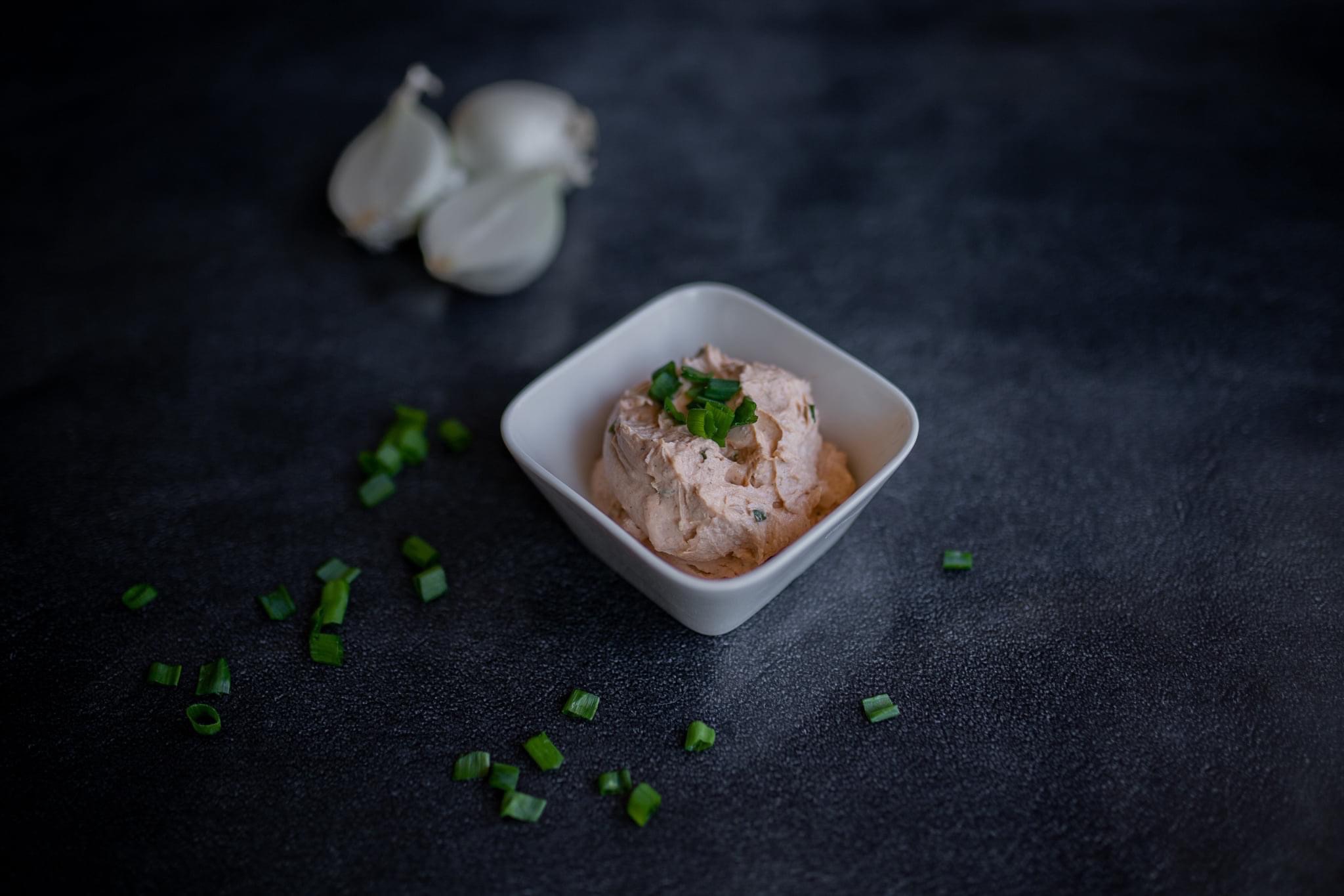 Kremowa pasta rybna w białej miseczce kwadratowej, posypana szczypiorkiem, obok dwie białe cebule i rozsypany szczypiorek na ciemnym tle.