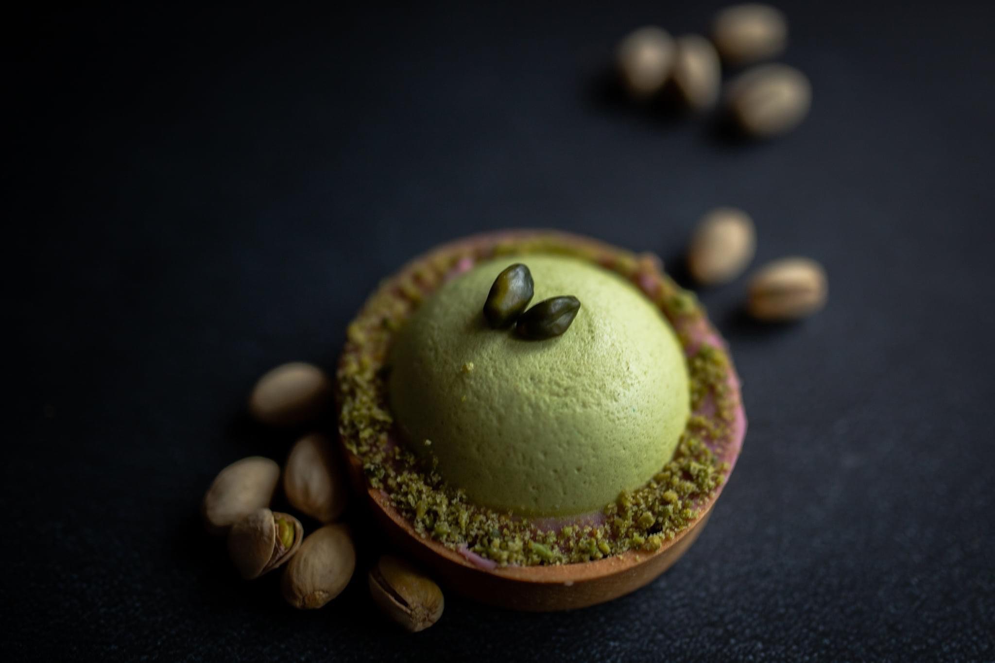Delikatna pistacjowa tarta z gładką kremową kopułą, posypana pistacjami i ozdobiona dwoma pistacjami na wierzchu, otoczona rozrzuconymi orzechami pistacjowymi na ciemnym tle.