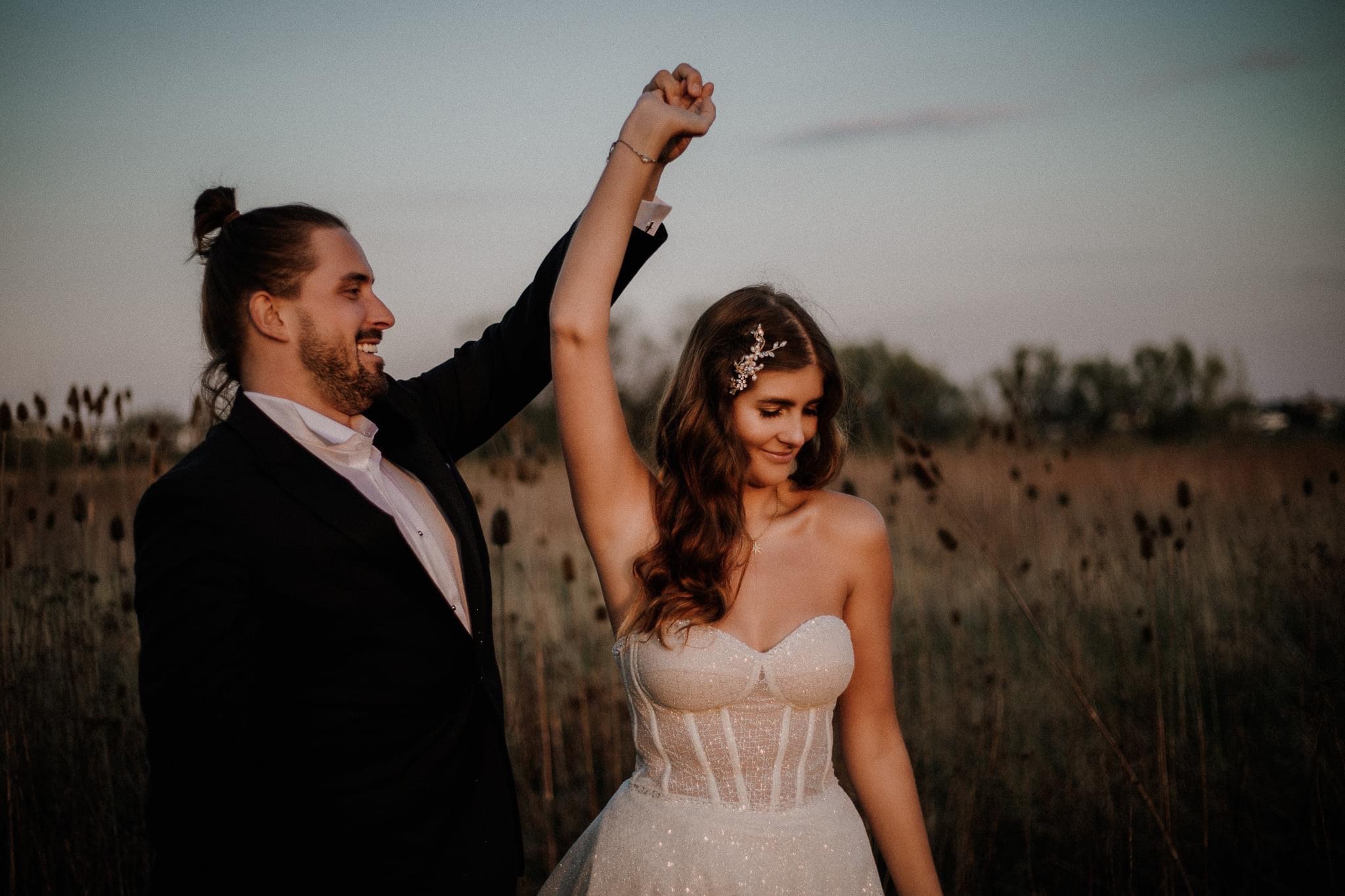 Para młoda trzymająca się za ręce na tle łąki w zachodzącym słońcu. Panna młoda w białej sukni bez ramiączek z błyszczącymi detalami i ozdobą we włosach. Pan młody w czarnym garniturze z rozpiętą...