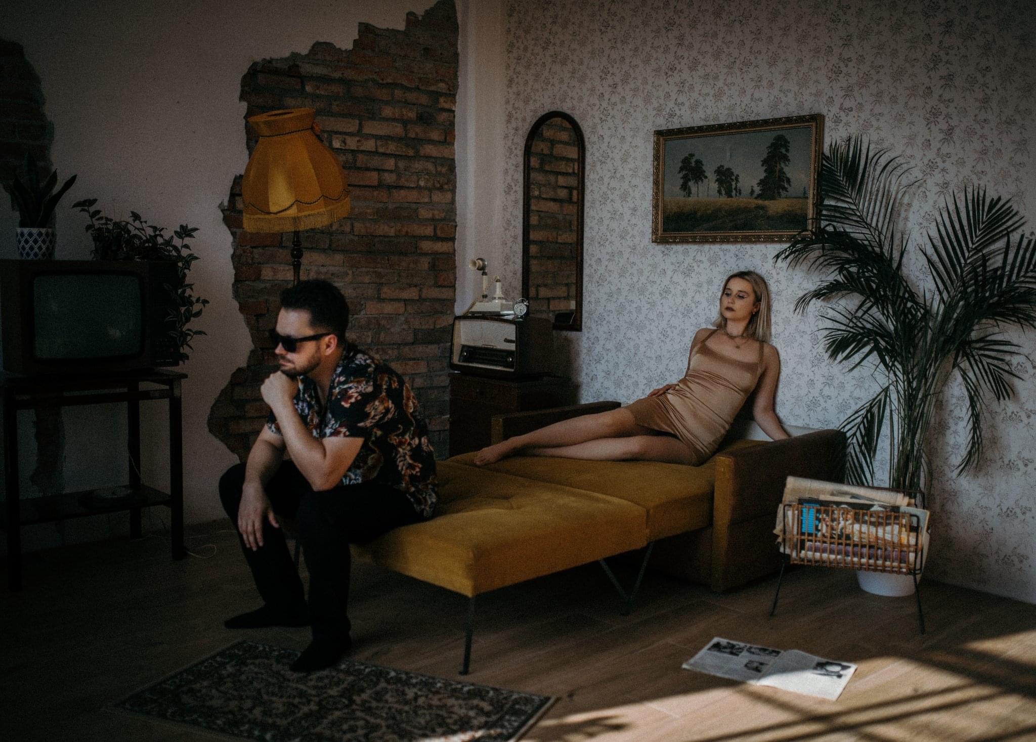 Para w stylizowanym wnętrzu retro: mężczyzna w kwiecistej koszuli siedzi zamyślony, a kobieta w złotej sukience leży na kanapie, otoczeni starodawnymi meblami i dekoracjami.