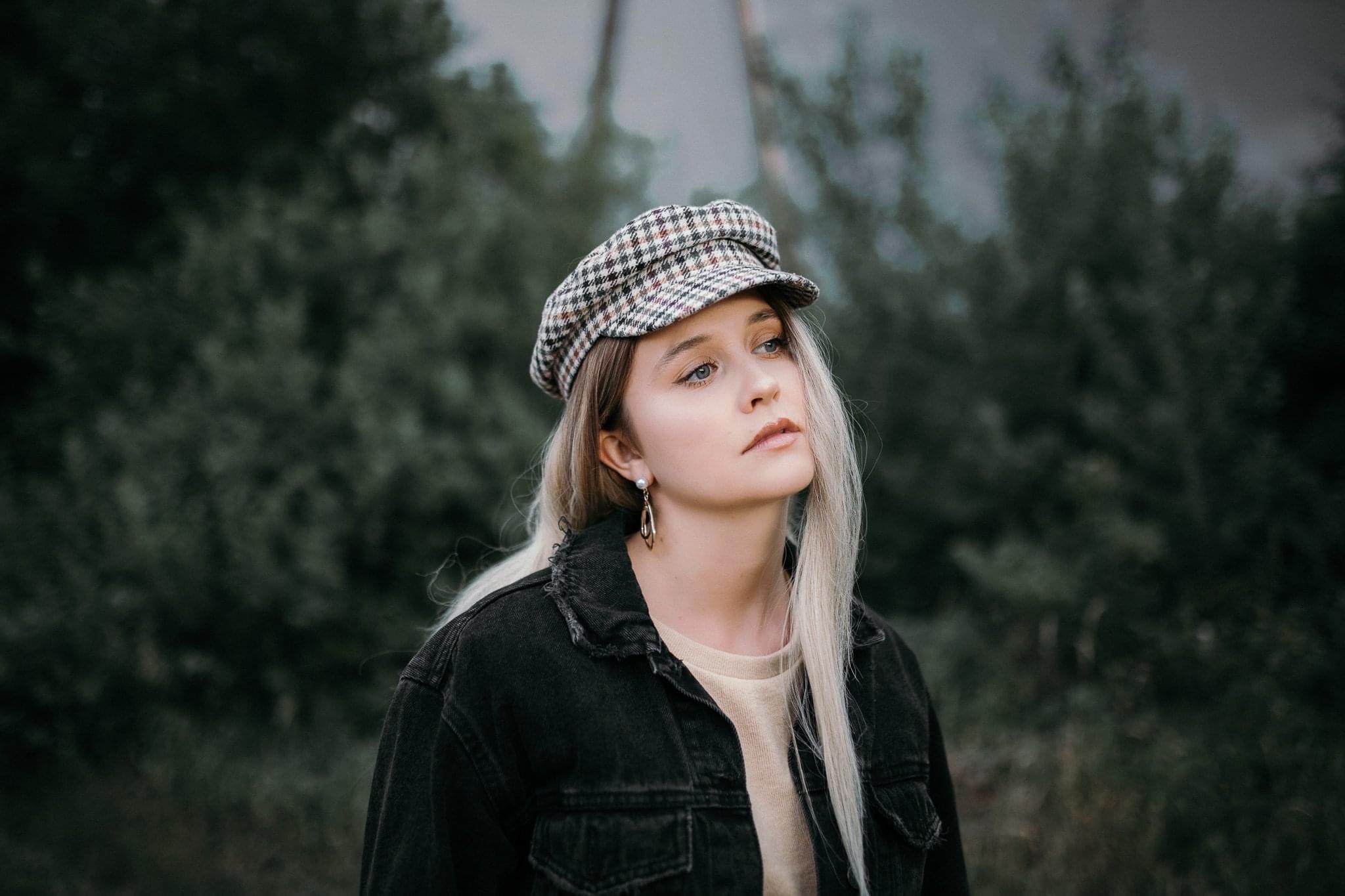 Portret młodej kobiety z blond włosami, w kraciastej czapce i czarnej kurtce jeansowej, z delikatnym makijażem i złotymi kolczykami na tle rozmytej zieleni.
