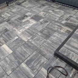 Ułożona, szara kostka brukowa na tarasie z ciemną obwódką i fragmentem narzędzia. Widok z góry, ukośny kadr.