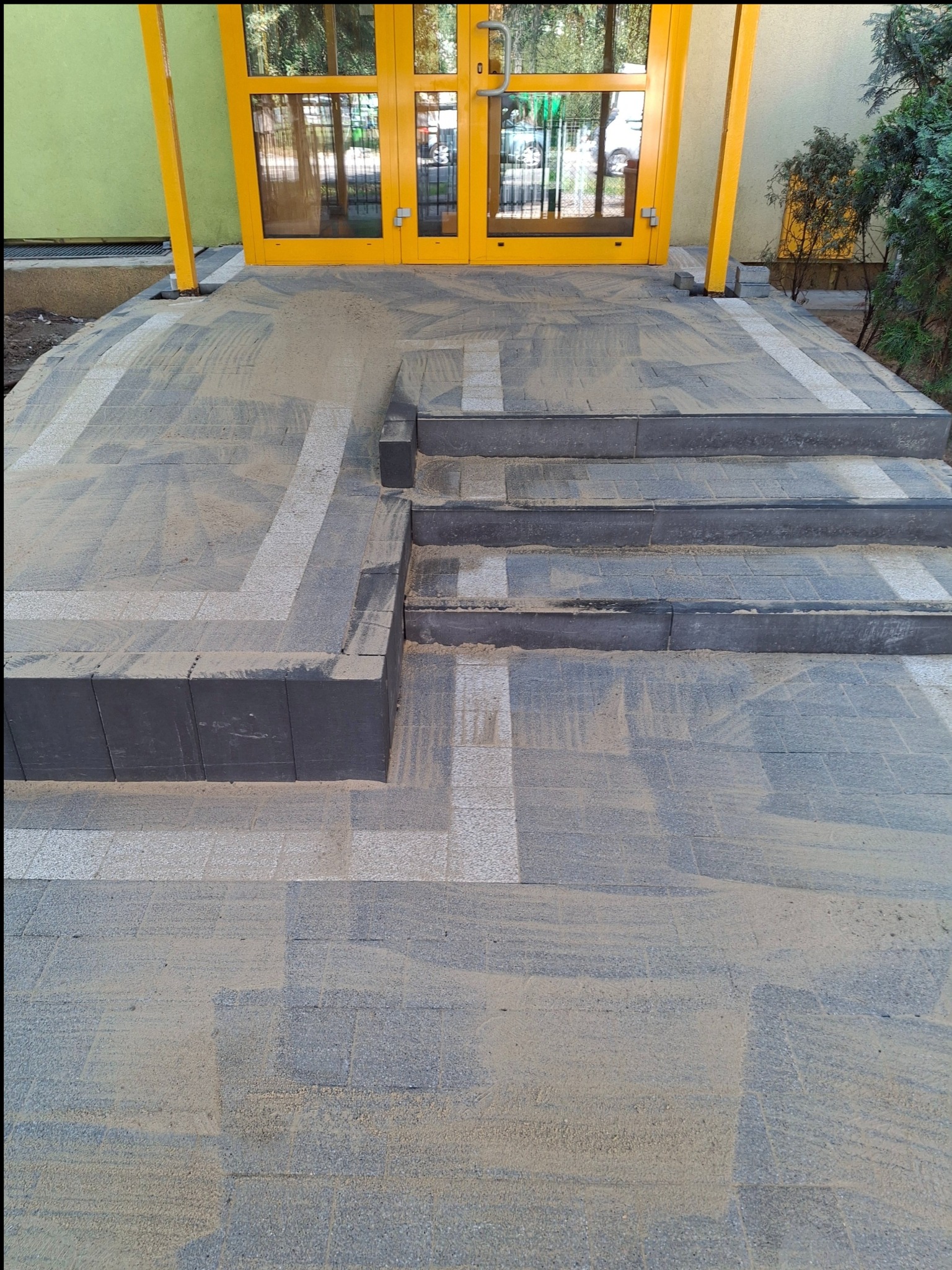 Nowo ułożona kostka brukowa w odcieniach szarości z białymi akcentami, prowadząca do żółtych drzwi wejściowych. Widoczne schody i ślady piasku na powierzchni.