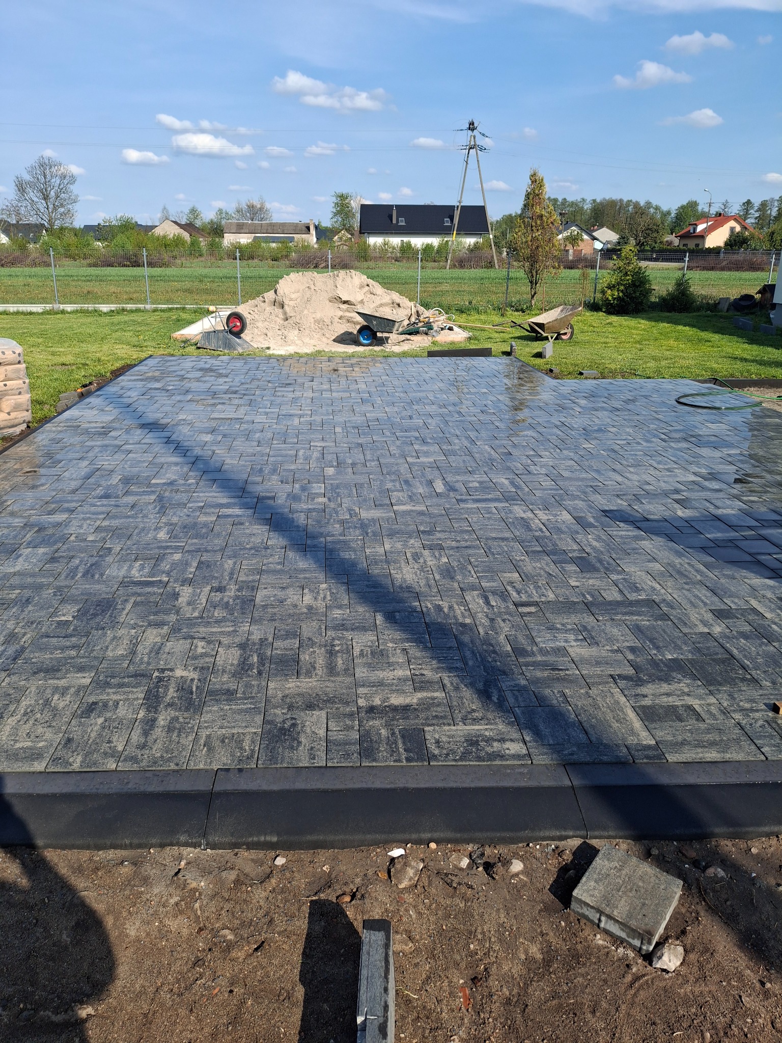 Ułożona, mokra kostka brukowa w Markach, z widocznym piaskiem i narzędziami w tle. Krawężnik oddziela kostkę od ziemi. Słoneczny dzień, niebieskie niebo.