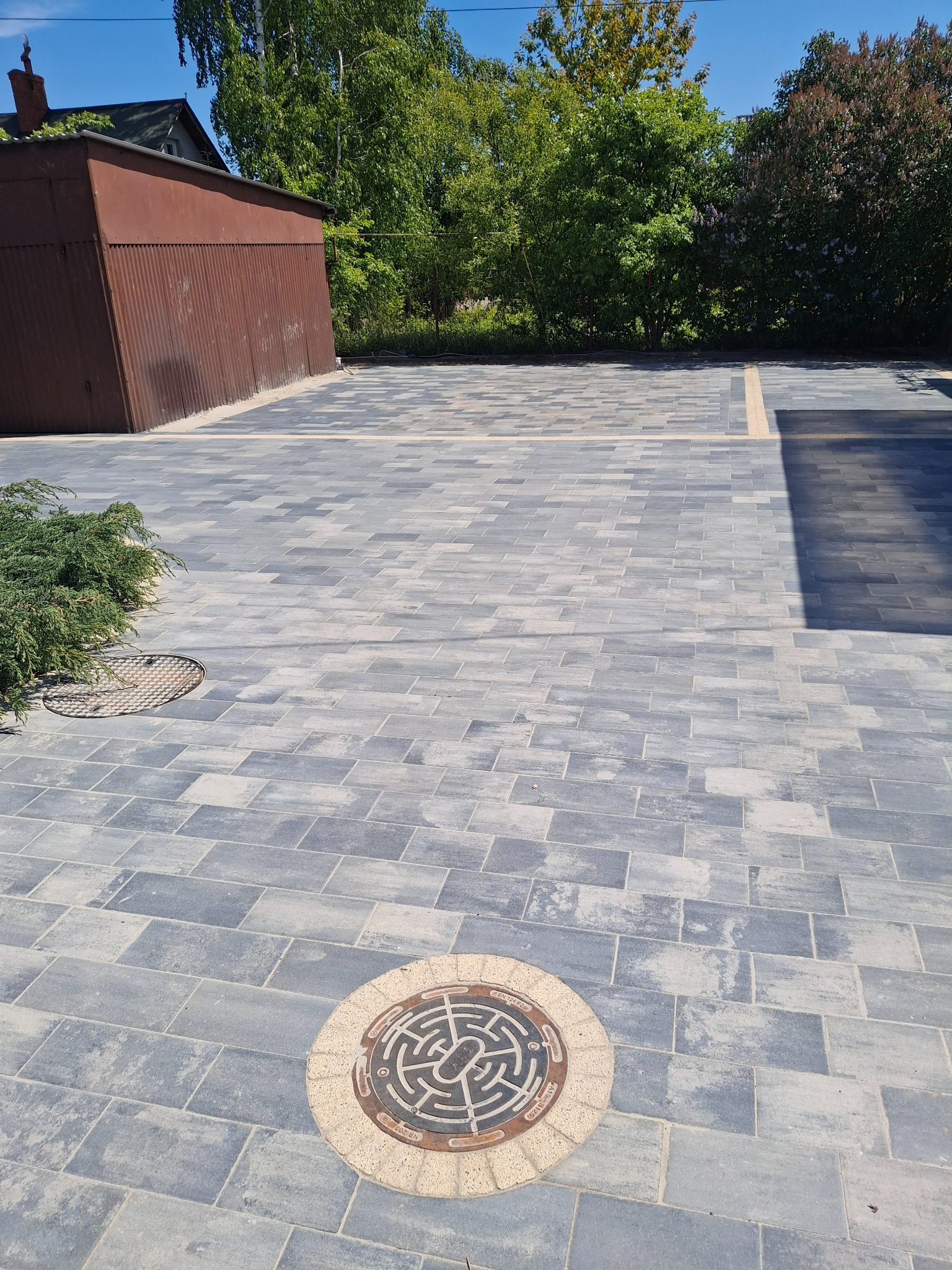 Ułożona, szara kostka brukowa tworząca duży plac z dwoma włazami kanalizacyjnymi. W tle blaszany garaż i zieleń drzew.