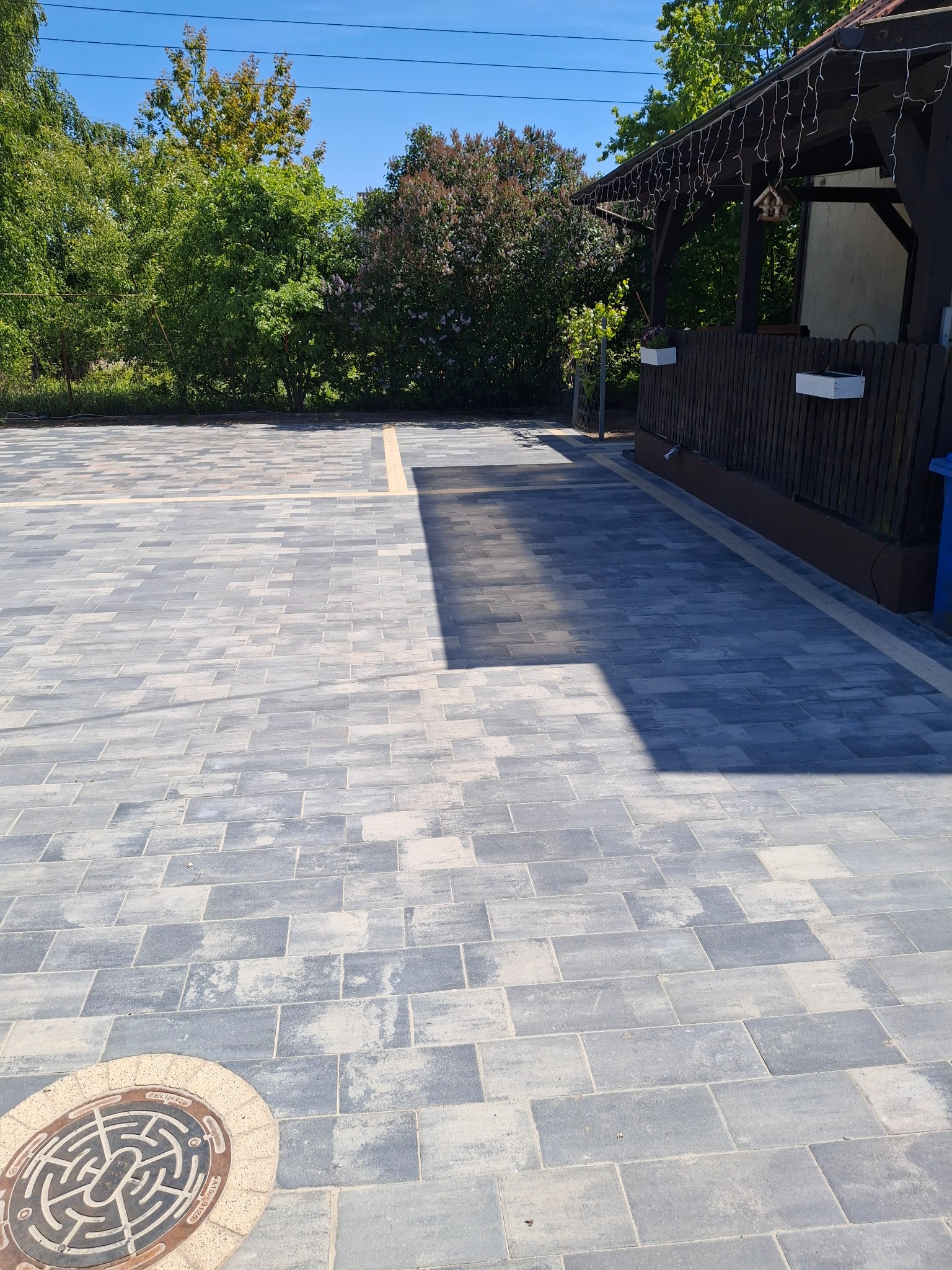 Szara kostka brukowa na podjeździe z żółtymi akcentami i studzienką kanalizacyjną. W tle zieleń drzew i fragment altany.