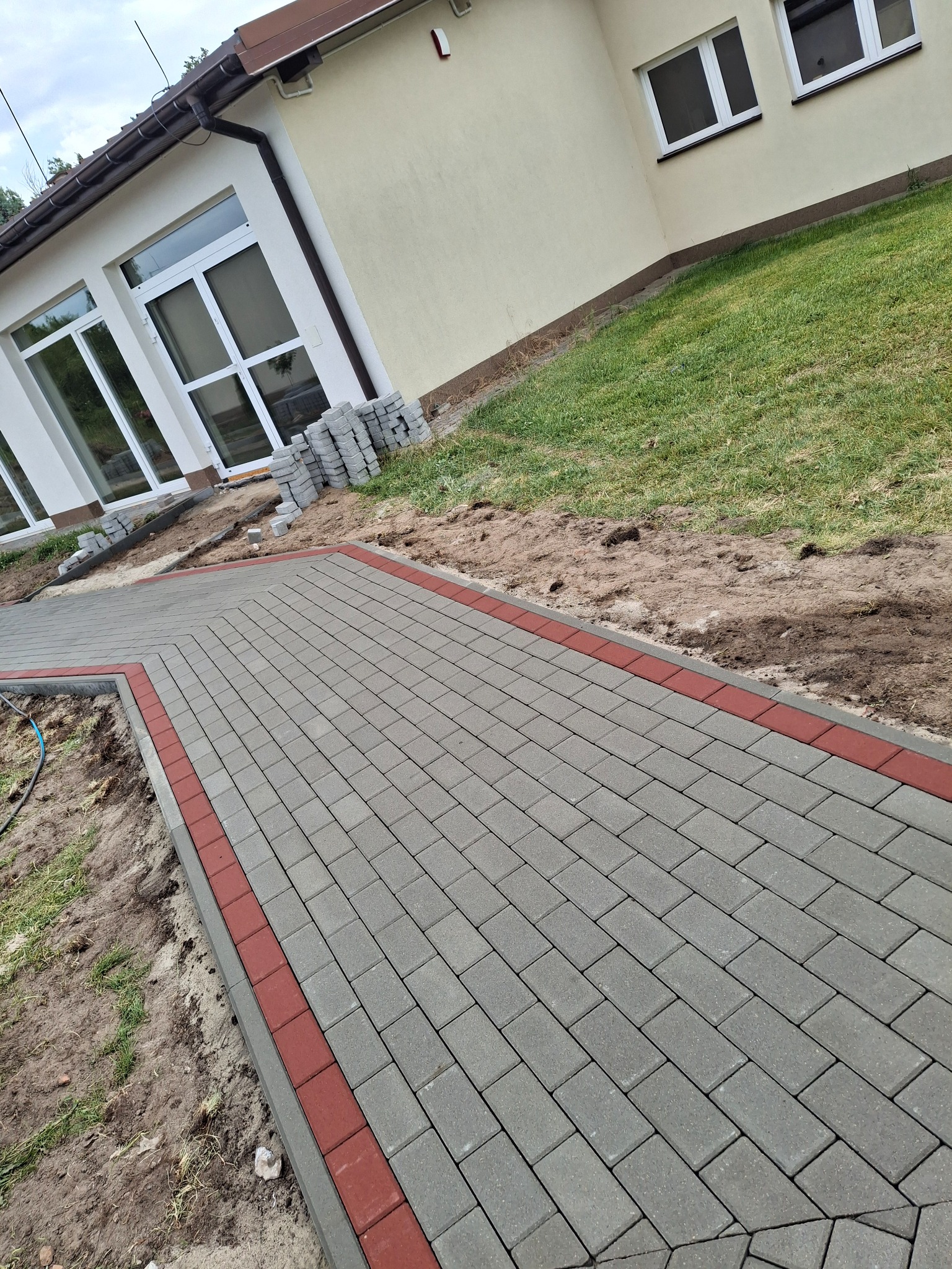 Ścieżka z kostki brukowej w odcieniach szarości z czerwonym obramowaniem, prowadząca wzdłuż budynku z białymi oknami. Część kostki jeszcze nieułożona.