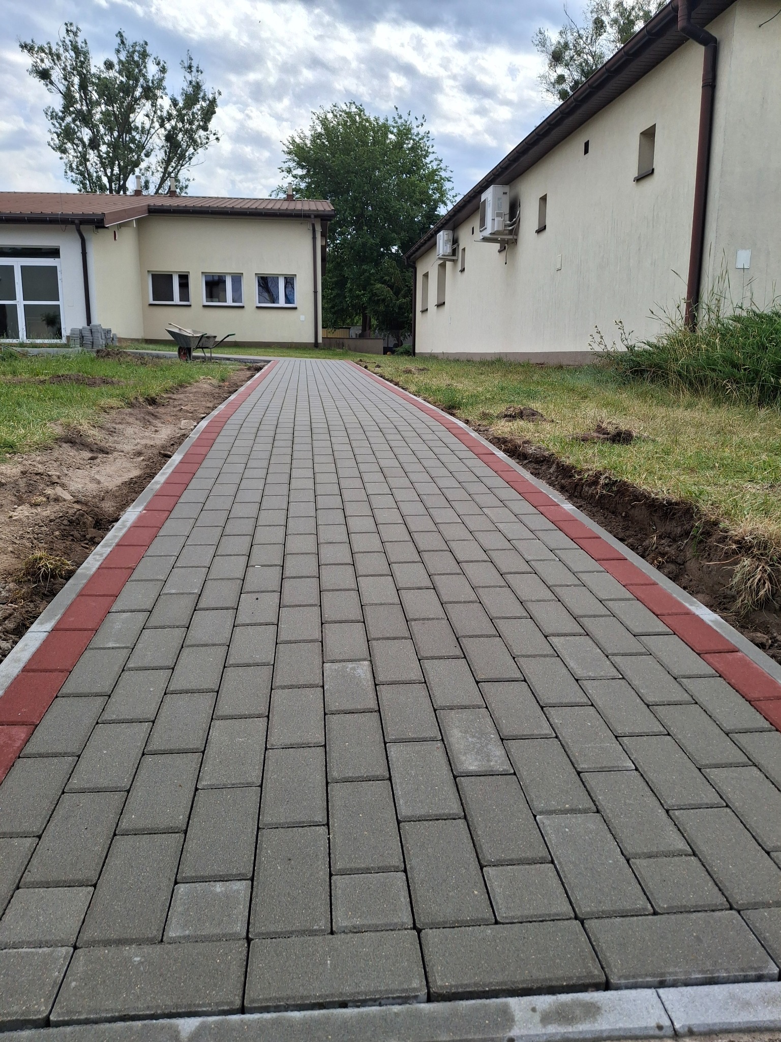 Nowa, długa ścieżka z szarej kostki brukowej z czerwonym obramowaniem, prowadząca między budynkami. W tle widoczny wózek budowlany i trawnik.