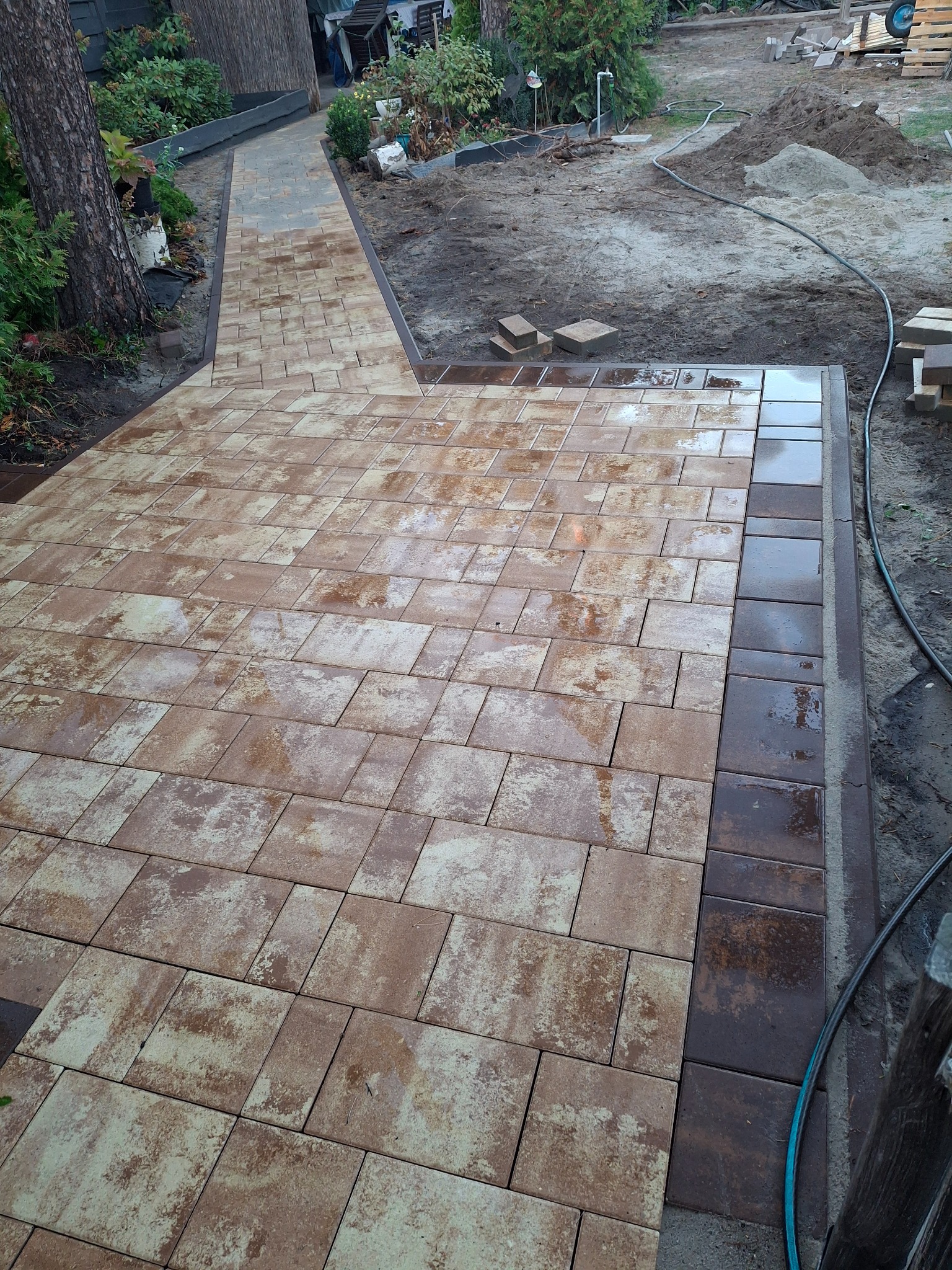Świeżo ułożona, brązowo-beżowa kostka brukowa na podwórku, z widocznym fragmentem ścieżki i przygotowanym terenem pod dalsze prace.
