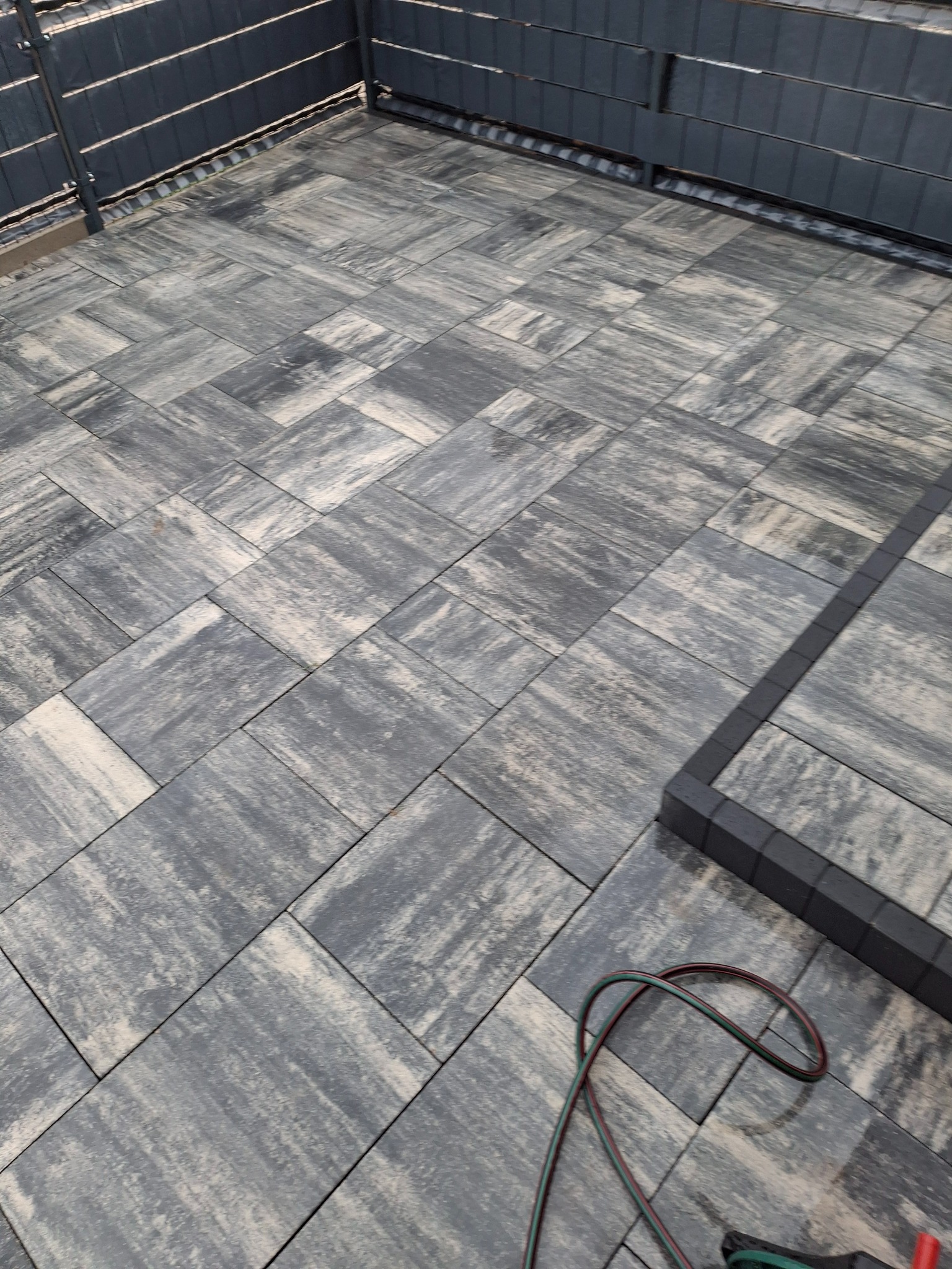 Ułożona, szara kostka brukowa na tarasie z ciemną obwódką i fragmentem narzędzia. Widok z góry, ukośny kadr.