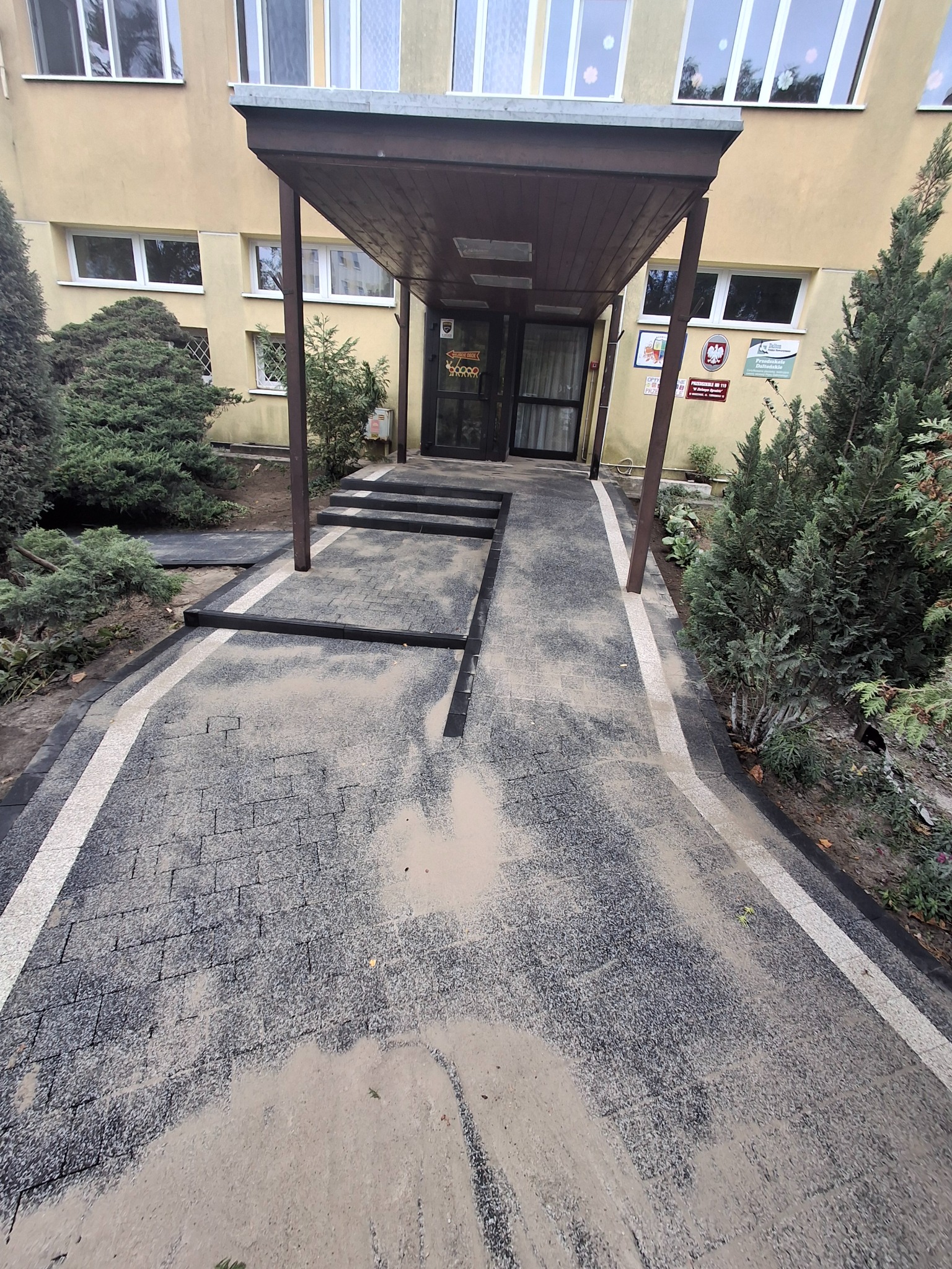 Nowo ułożona, szara kostka brukowa z białymi obrzeżami prowadząca do wejścia budynku. Widoczne elementy konstrukcyjne i pozostałości piasku.