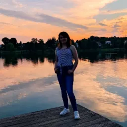 Kobieta w paski stoi na drewnianym pomoście nad jeziorem o zachodzie słońca, z odbiciem chmur w wodzie.