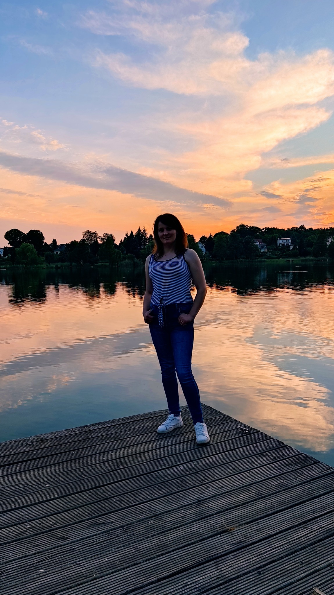 Kobieta w paski stoi na drewnianym pomoście nad jeziorem o zachodzie słońca, z odbiciem chmur w wodzie.