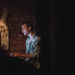 Młody mężczyzna w koszuli w kwiaty gra na pianinie na tle ceglanej ściany z metalową dekoracją w kształcie litery K.