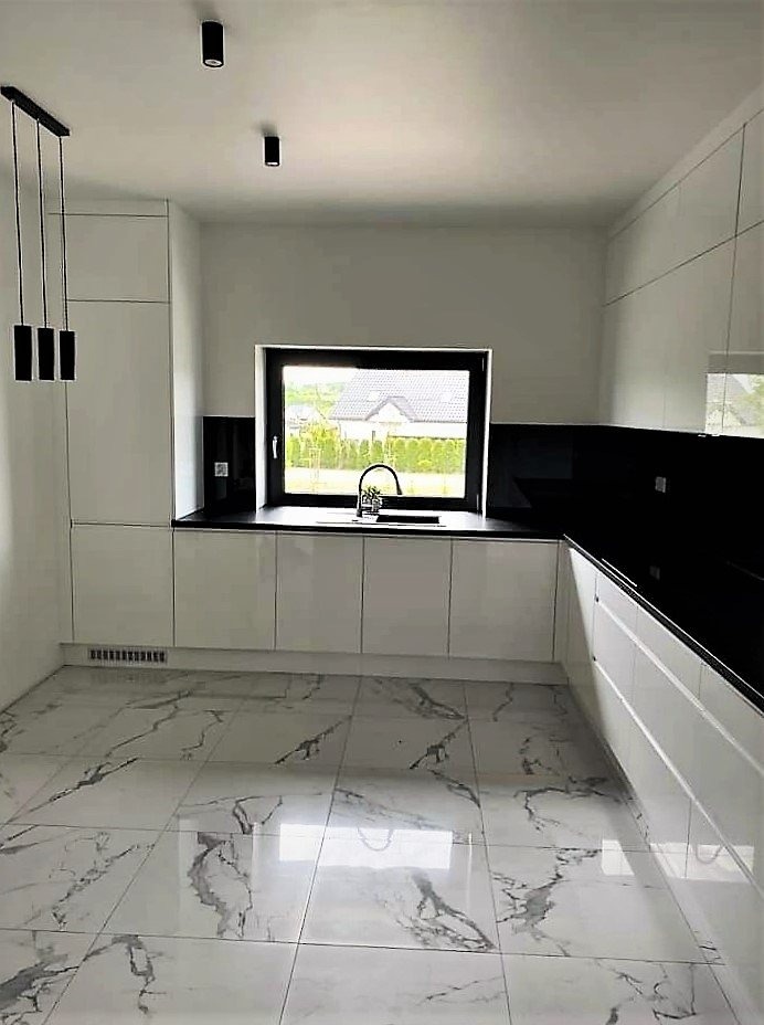 Nowoczesna kuchnia z białymi, lakierowanymi frontami, czarnym blatem i panelem nad blatem, marmurową podłogą oraz oknem z widokiem na ogród.