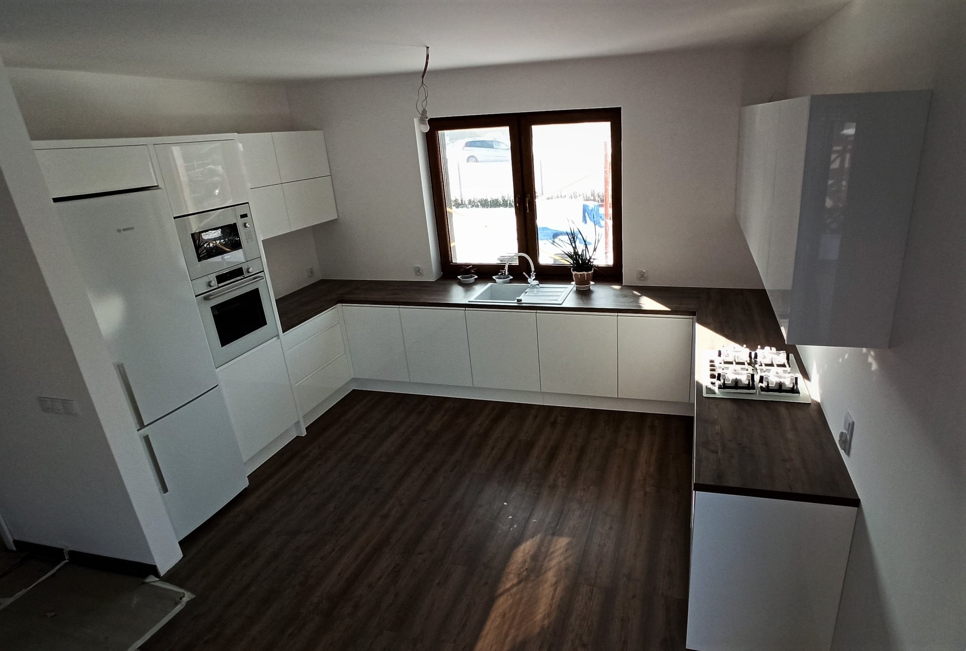 Nowoczesna, jasna kuchnia z białymi frontami i drewnianym blatem, widok na okno z rośliną doniczkową, brązowa podłoga, wbudowany sprzęt AGD.