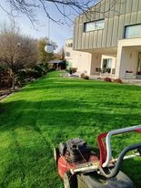 Świeżo skoszony trawnik przed nowoczesnym domem, widoczne ślady po kosiarce, na pierwszym planie fragment czerwonej kosiarki, w tle bujna zieleń i architektoniczne detale budynku.